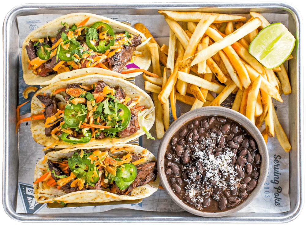 Three grilled steak tacos topped with jalapeños, cilantro, crispy onions, shredded vegetables and spicy sauce, served with fries, black beans and lime.