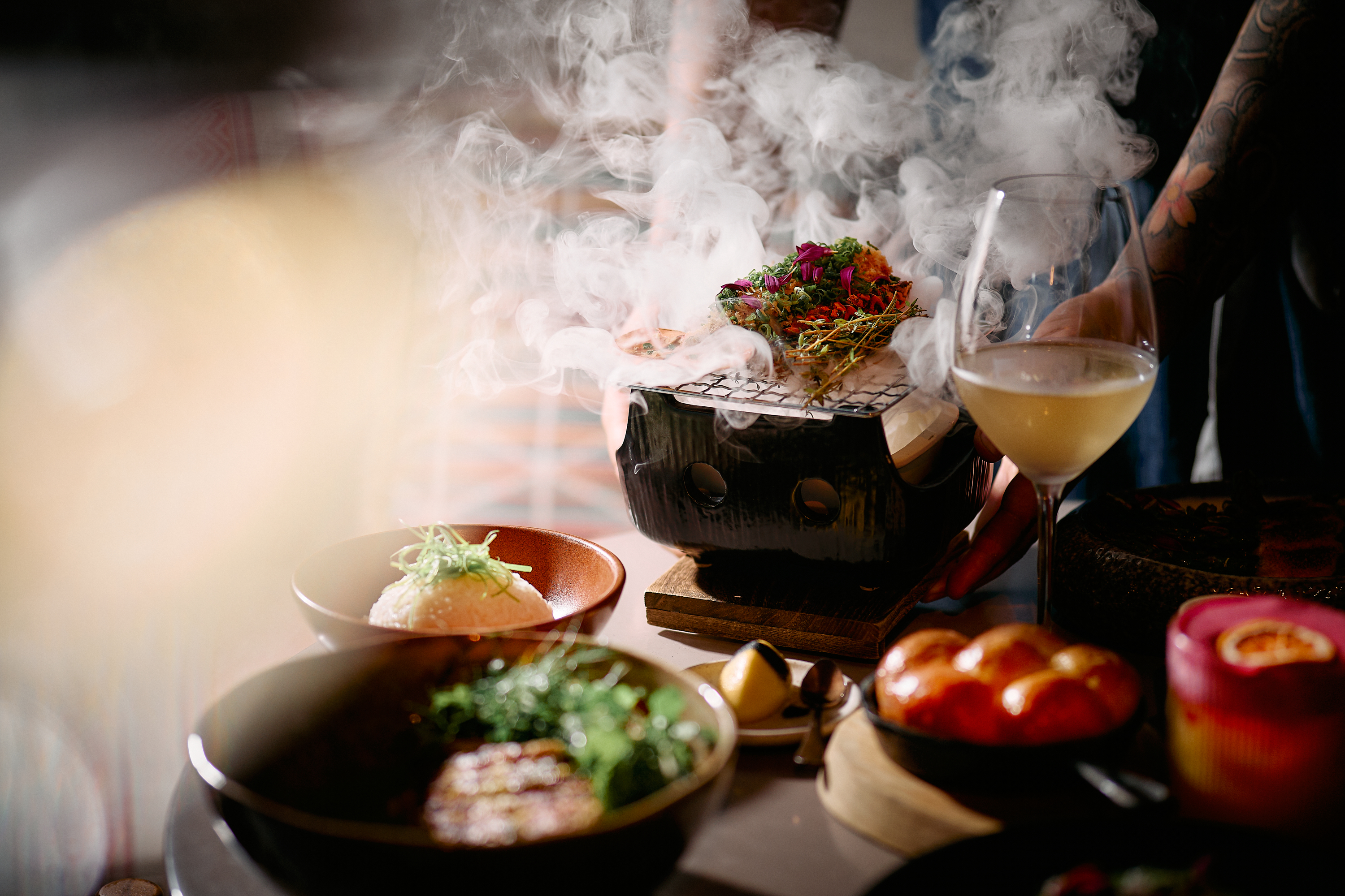 Plated dishes surround a small tabletop grill releasing dense smoke, with colorful garnish on top and a glass of light-colored beverage nearby