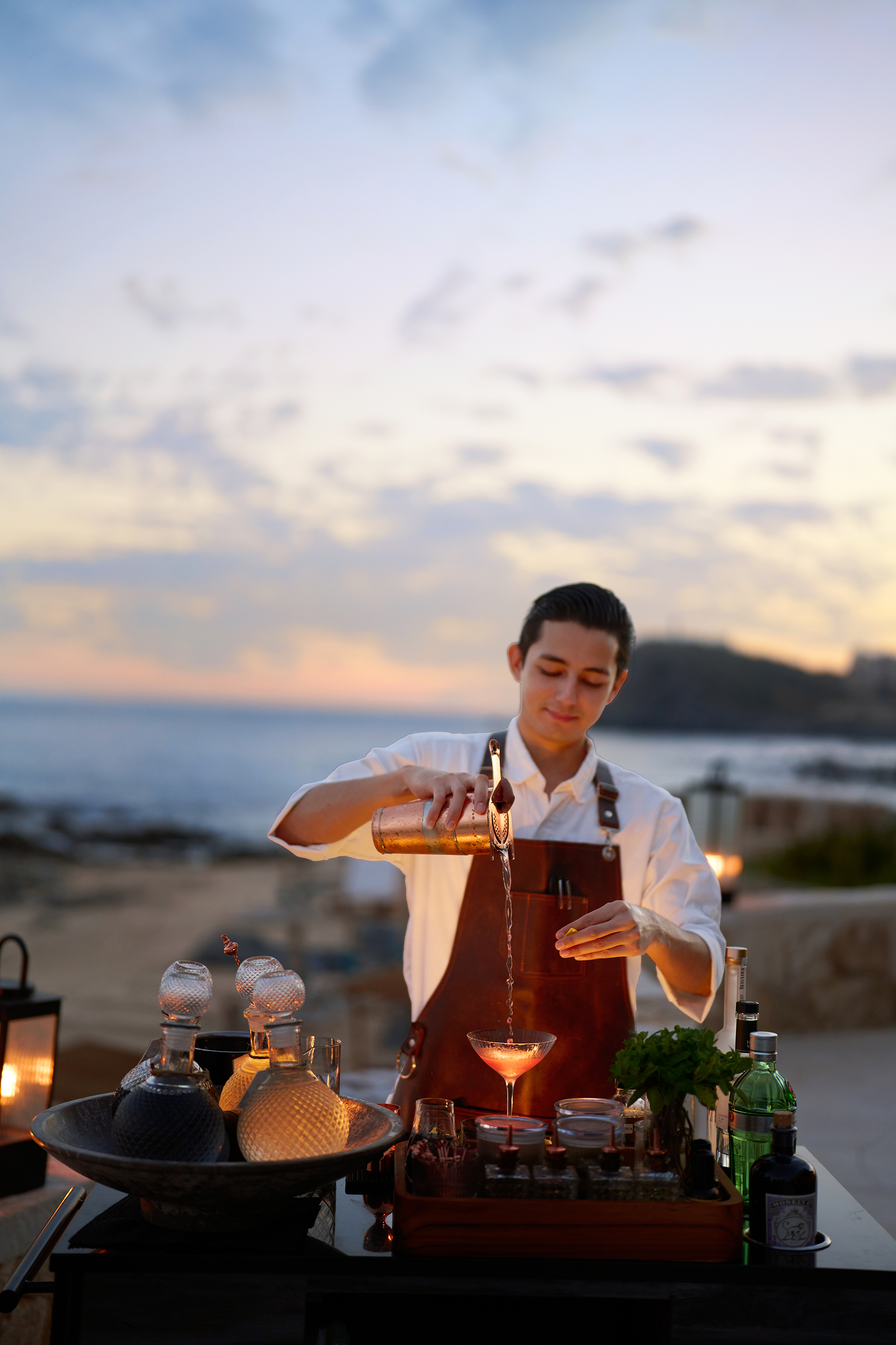Bartender preparing a handcrafted cocktail at Cayao’s oceanfront outdoor bar at sunset, surrounded by premium spirits, fresh garnishes, and coastal views.