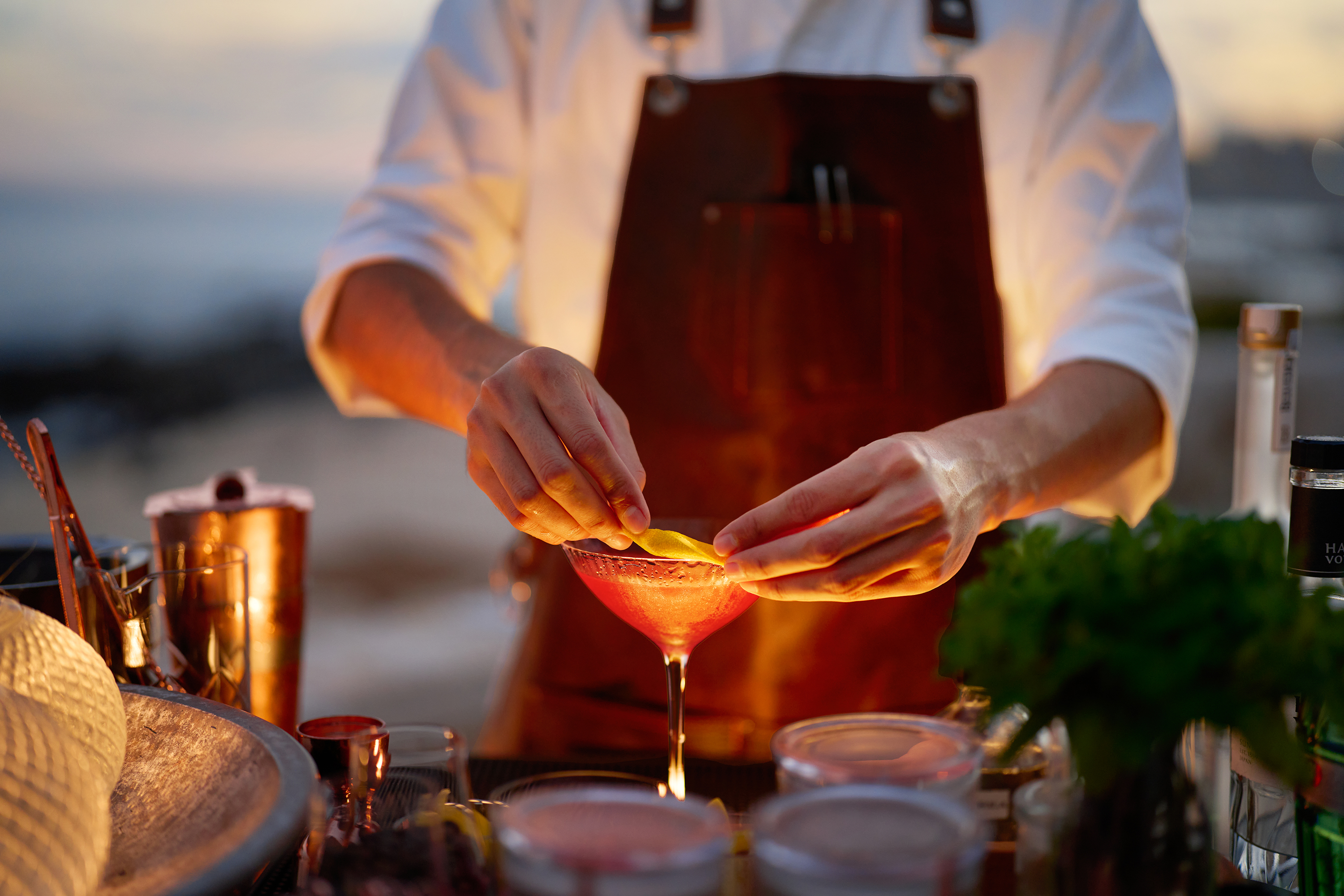 Bartender garnishing a pink cocktail with a citrus peel at an outdoor bar during sunset at Cayao, with fresh herbs and cocktail ingredients in the foreground.