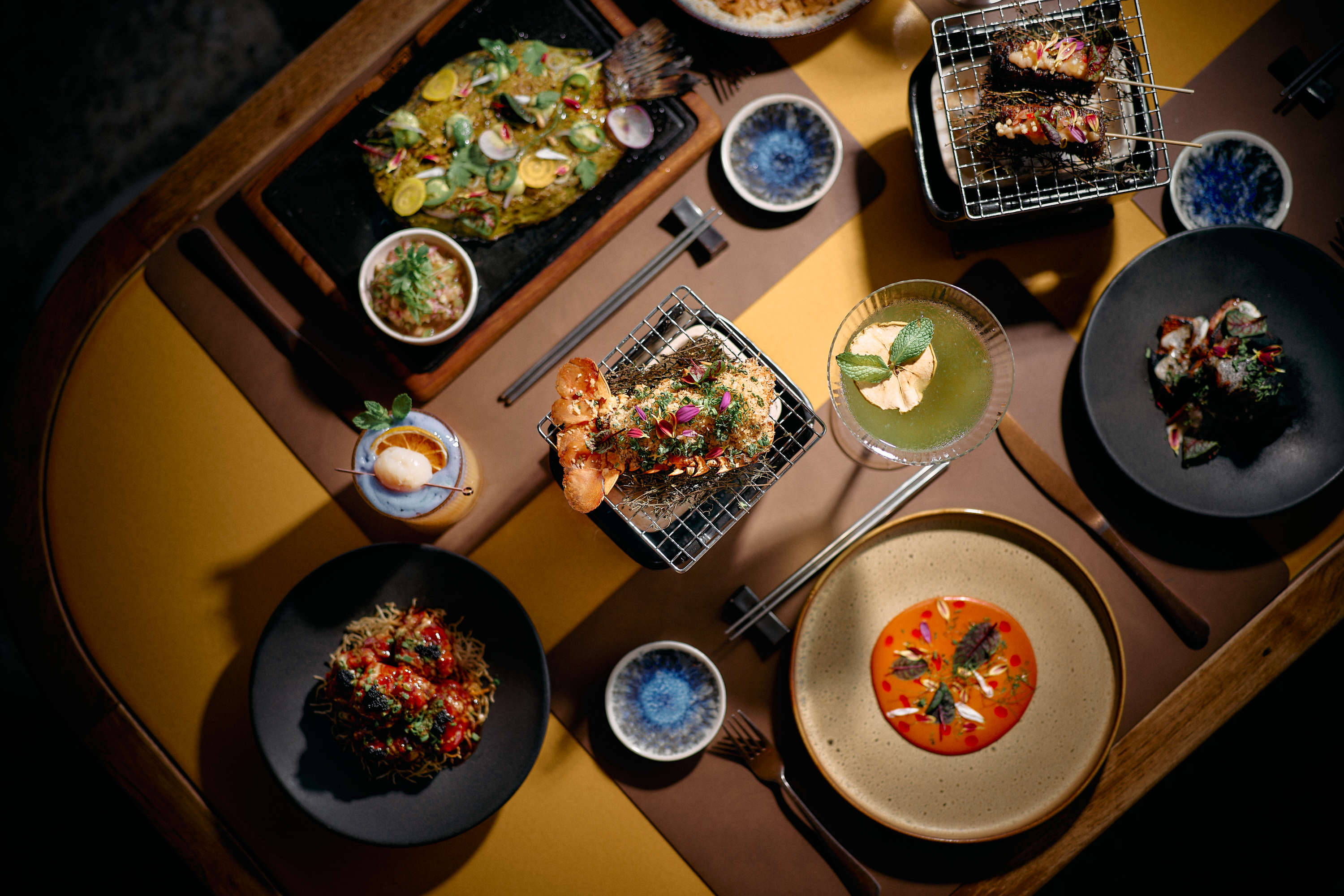 Assorted plated dishes arranged on a table, including grilled items on small metal racks, vibrant soups, and garnished seafood, viewed from above