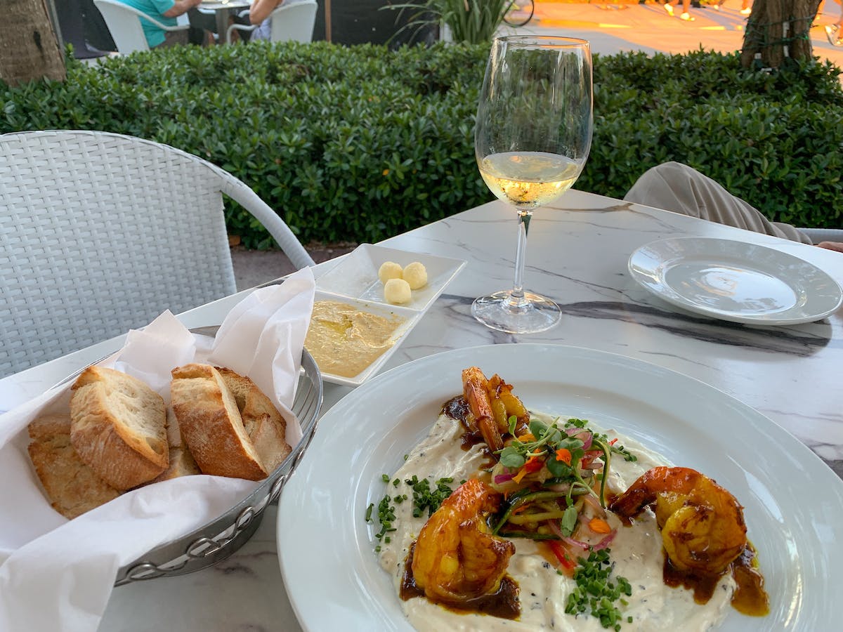 a table setting with a plate of shrimp, a bowl with bread, butter on the side and a white wine glass