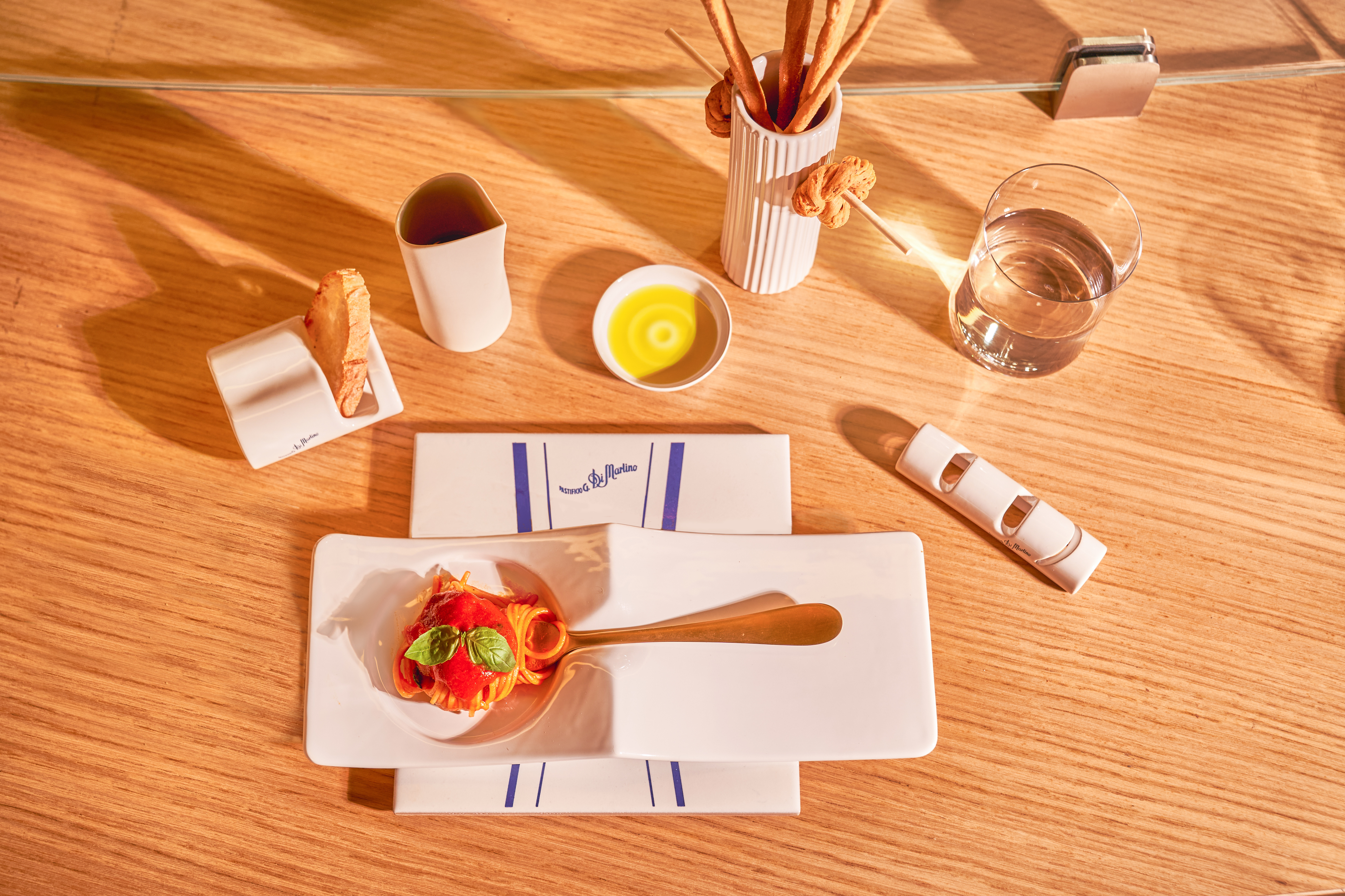 Overhead view of a set table with a small plate of pasta, ceramic condiment dishes, a glass of water, on light wood