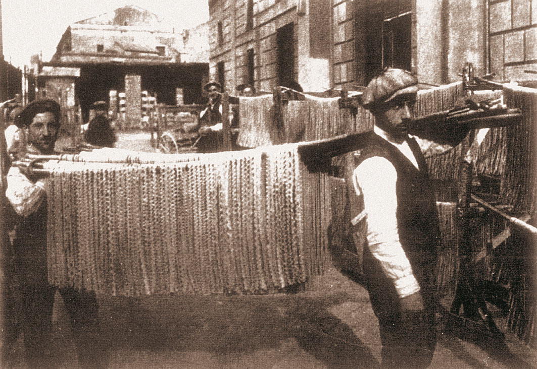 Black-and-white archival photo of a worker in Gragnano carrying a rack of long pasta strands drying in the street