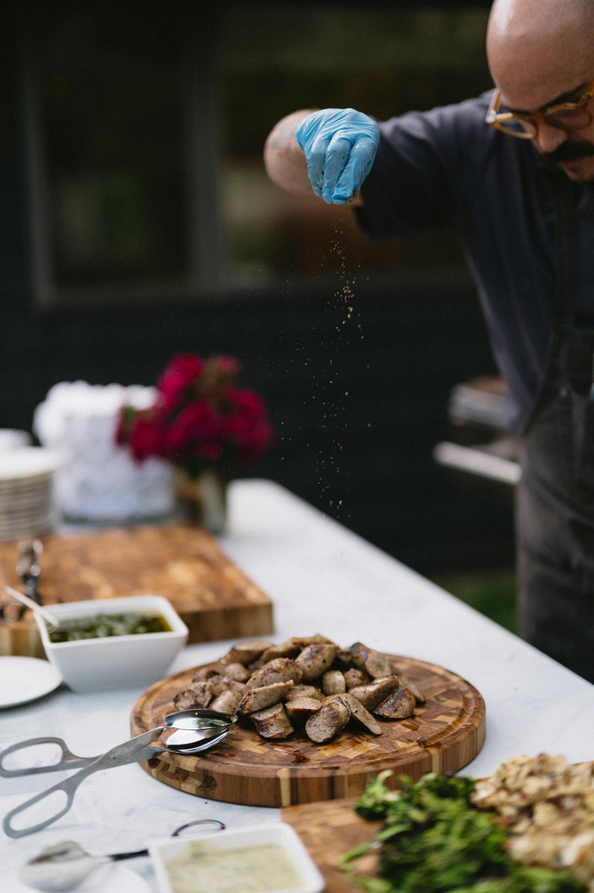 A chef from Michelle Bernstein Catering sprinkling salt and seasoning on a plate of sausages.