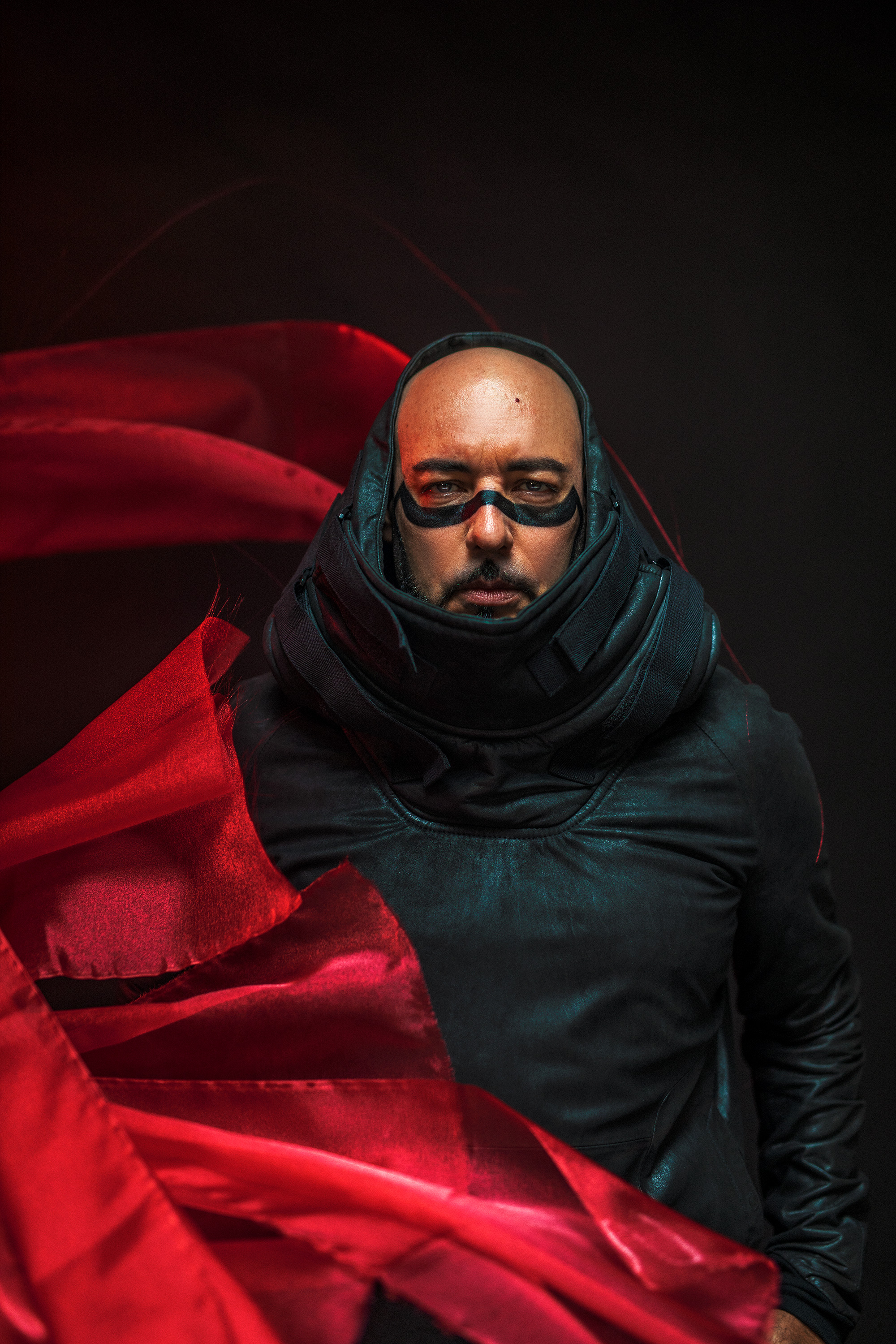 Dramatic portrait of a man in black futuristic attire with eye paint and flowing red fabric