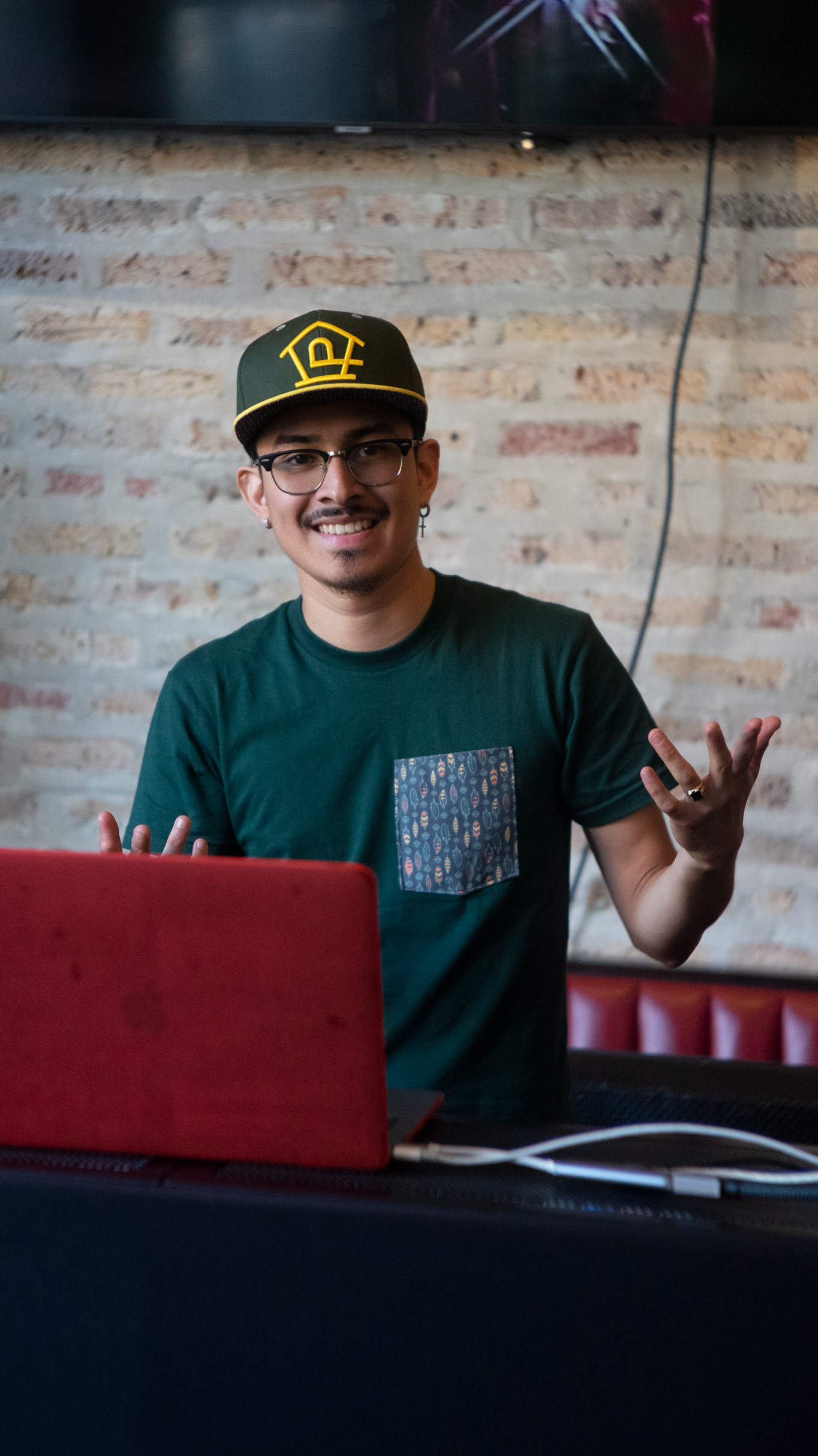 Young man wearing a tshirt and hat from The Perch standing at his DJ booth behind a laptop