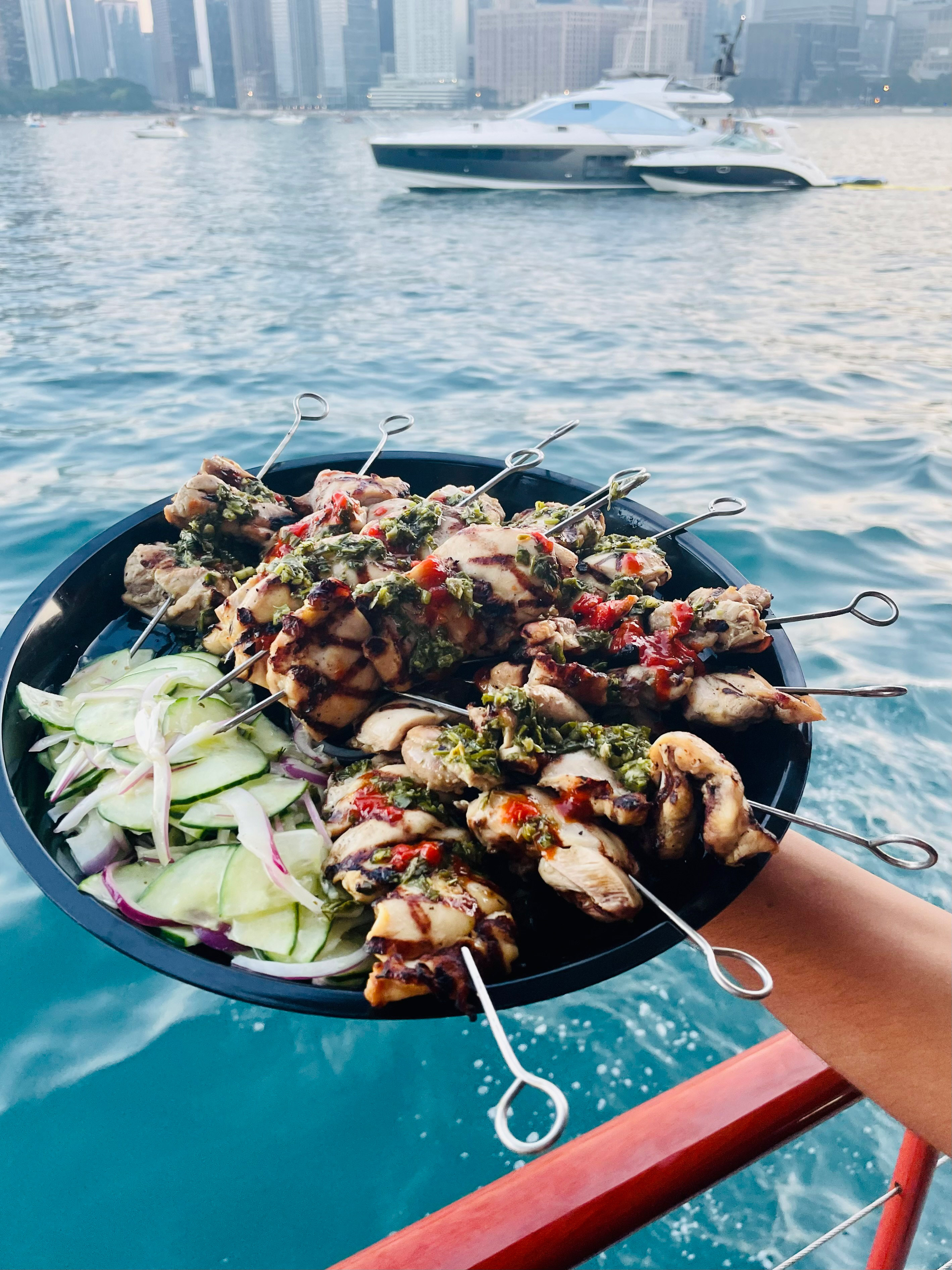 A plate of chicken schwarma skewers being held with Lake Michigan, a boat, and the city skyline visible in the background