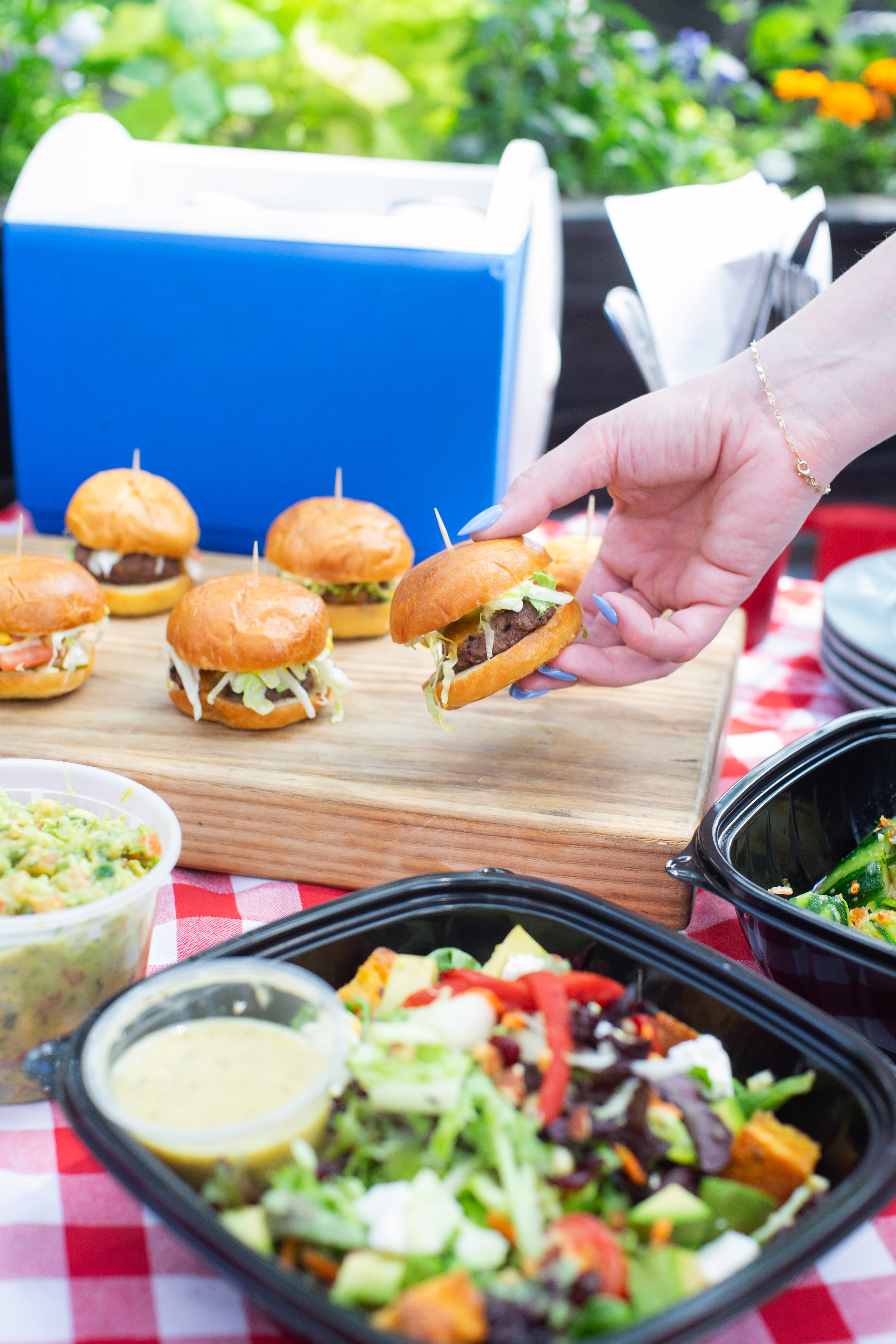 A hand reaching towards a picnic table set with sliders, salads and other bites