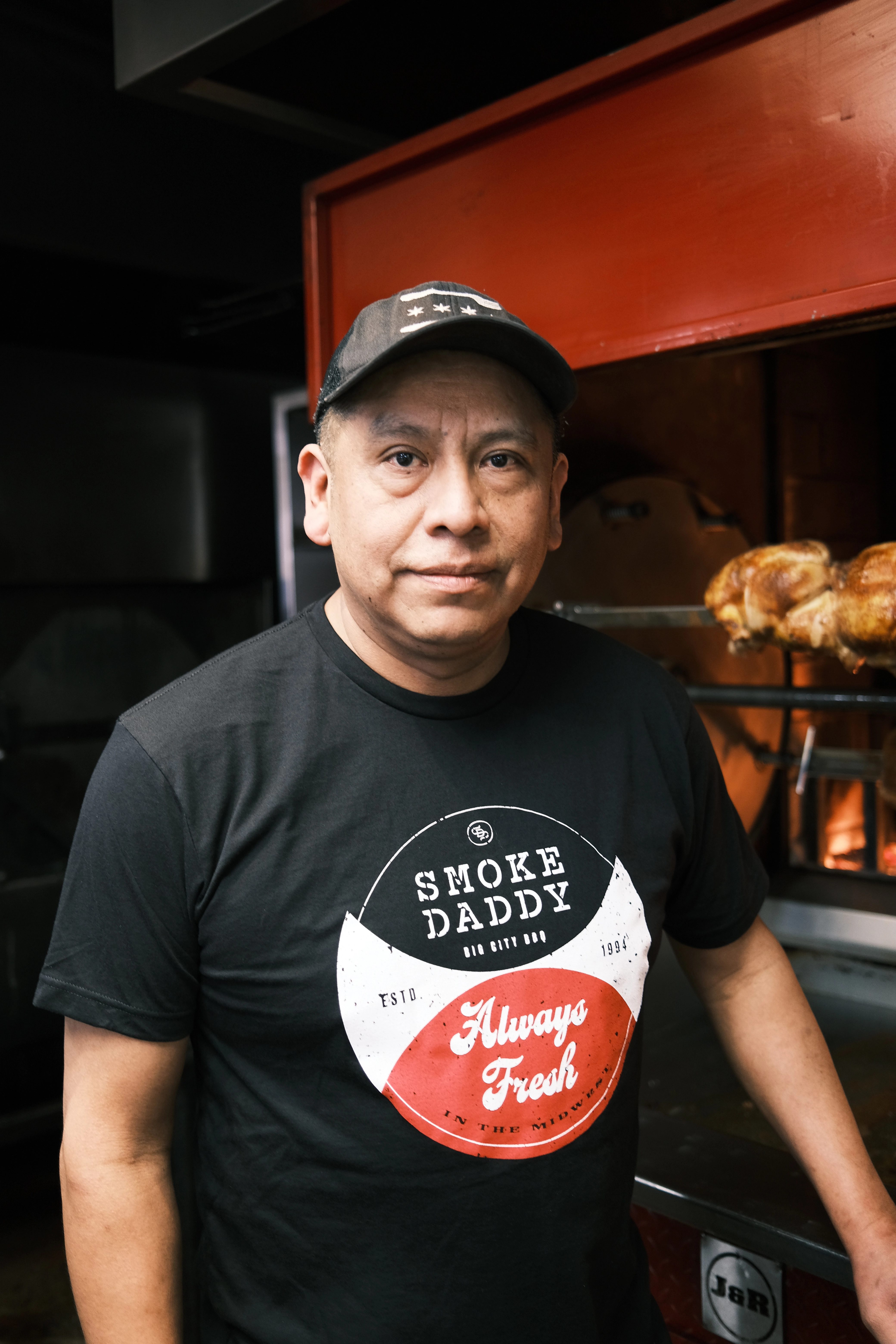 Man standing in front of a rotisserie and smoker wearing a Smoke Daddy tshirt