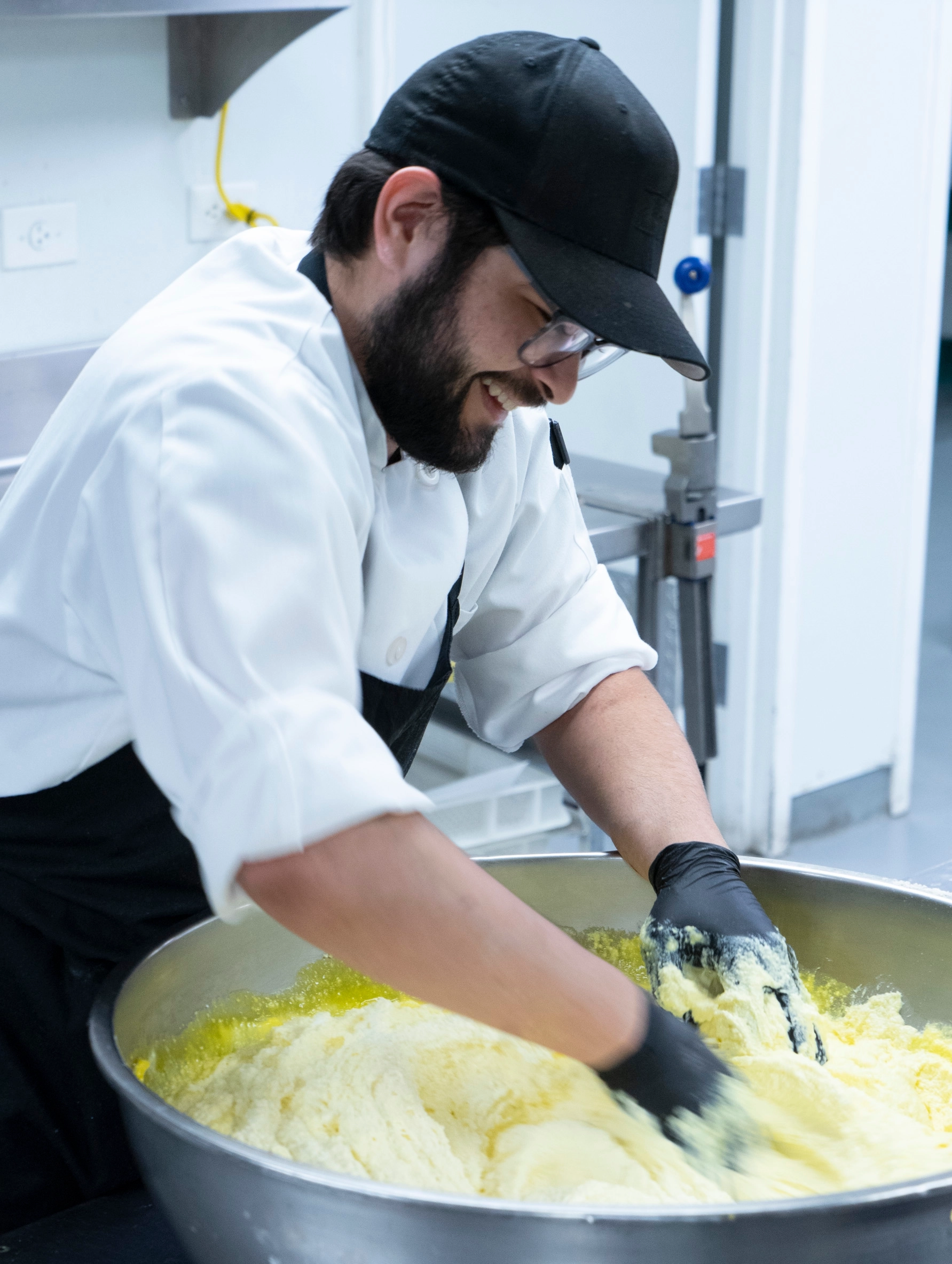 man in chef coat mixing eggs and flour in a massive silver bowl