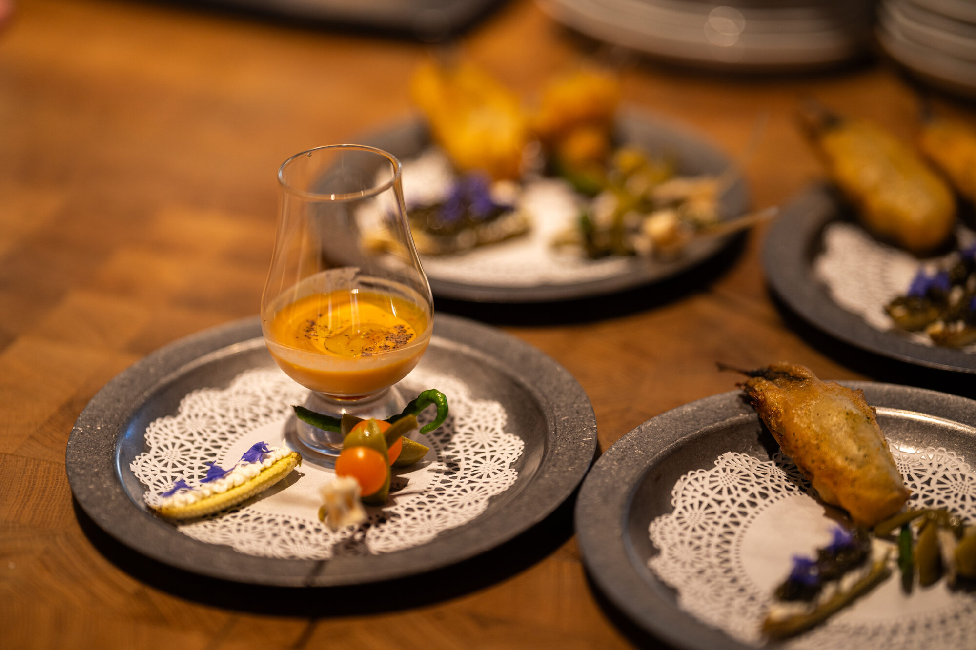An opening course of snacks on pewter plates with paper doilies, including a chilled soup poured into a tasting glass