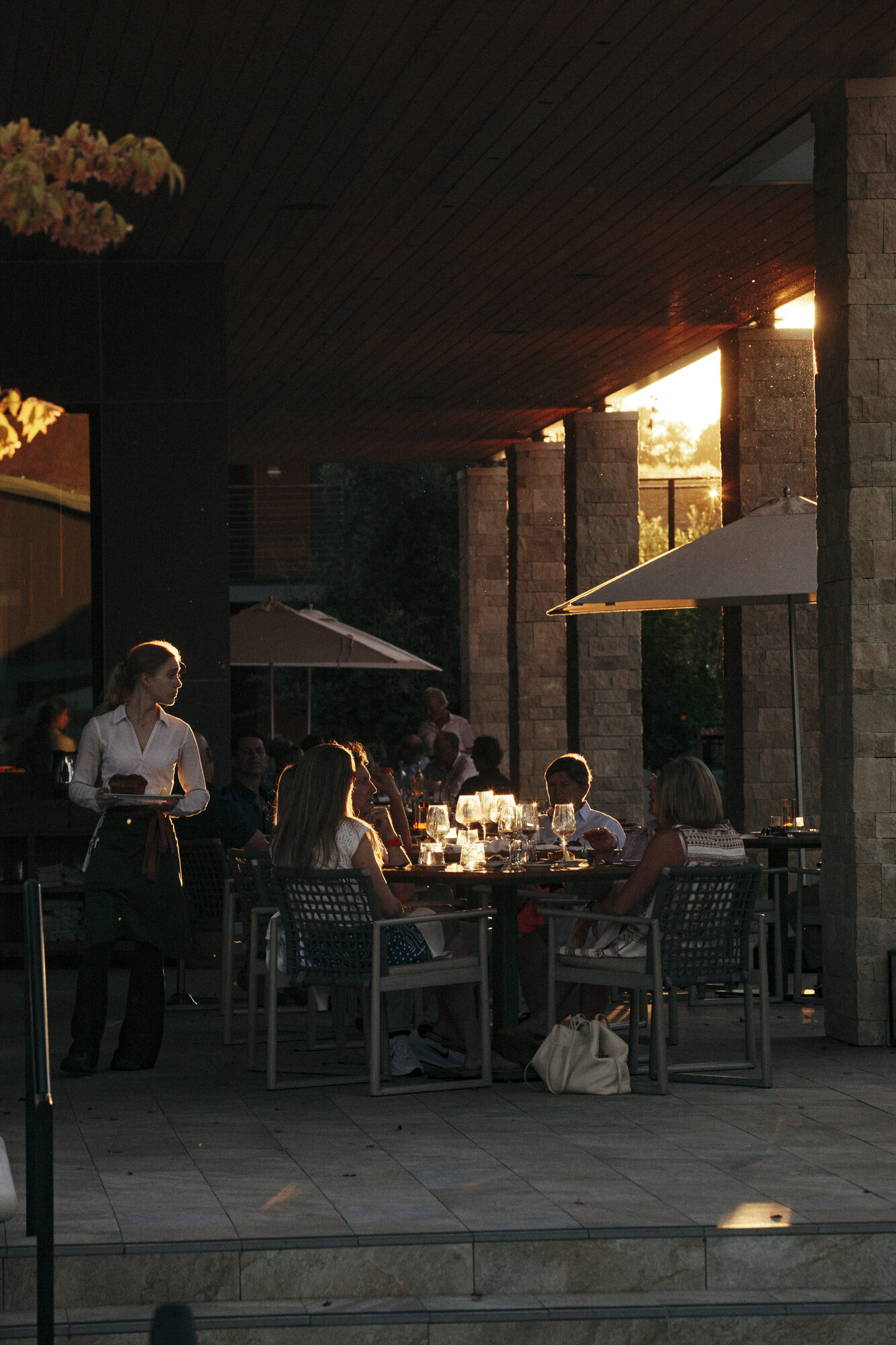 Guests dining on the Folia patio at golden hour as a server carries a tray to the table
