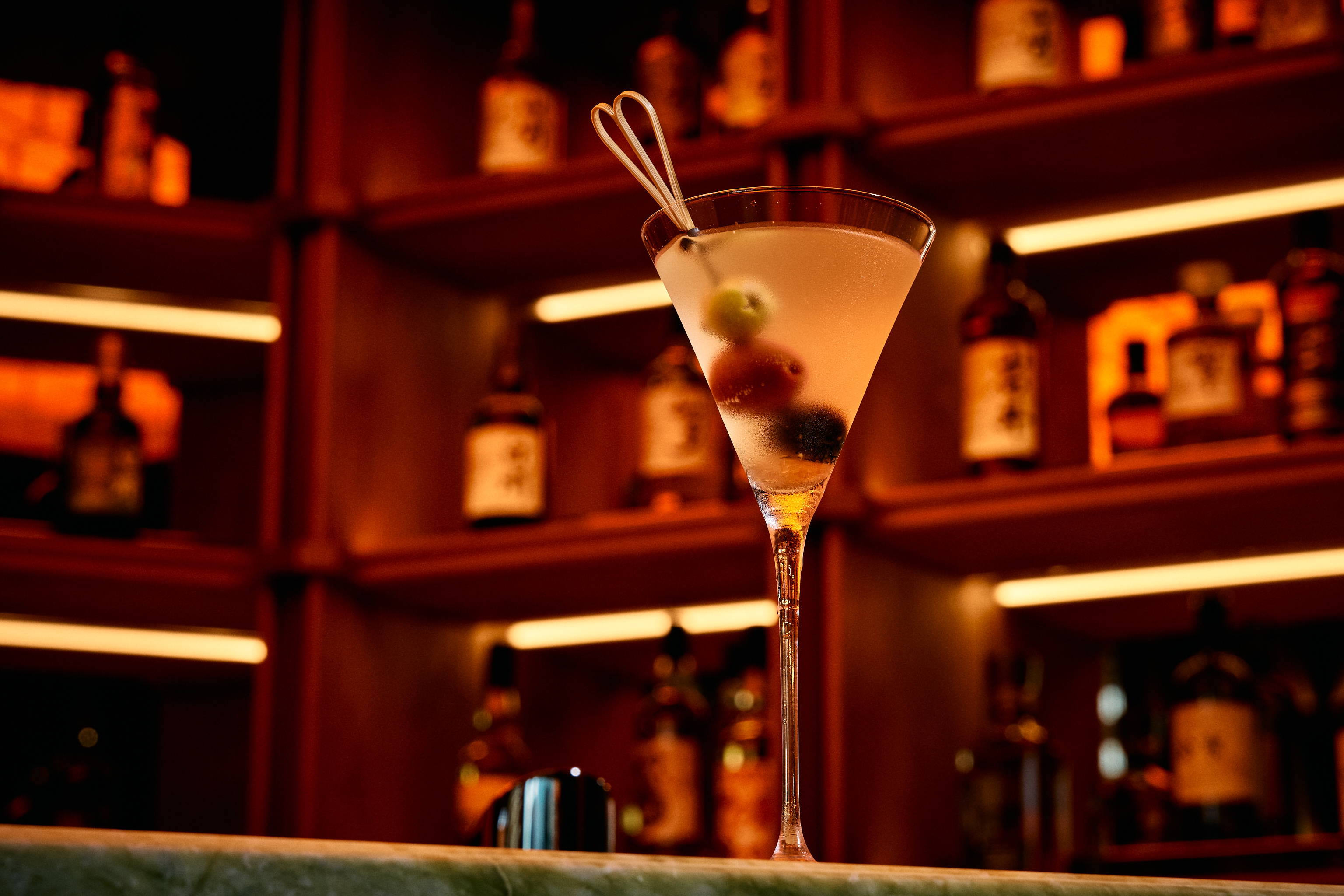 a martini glass with liquid and olives in front of shelves of bottles