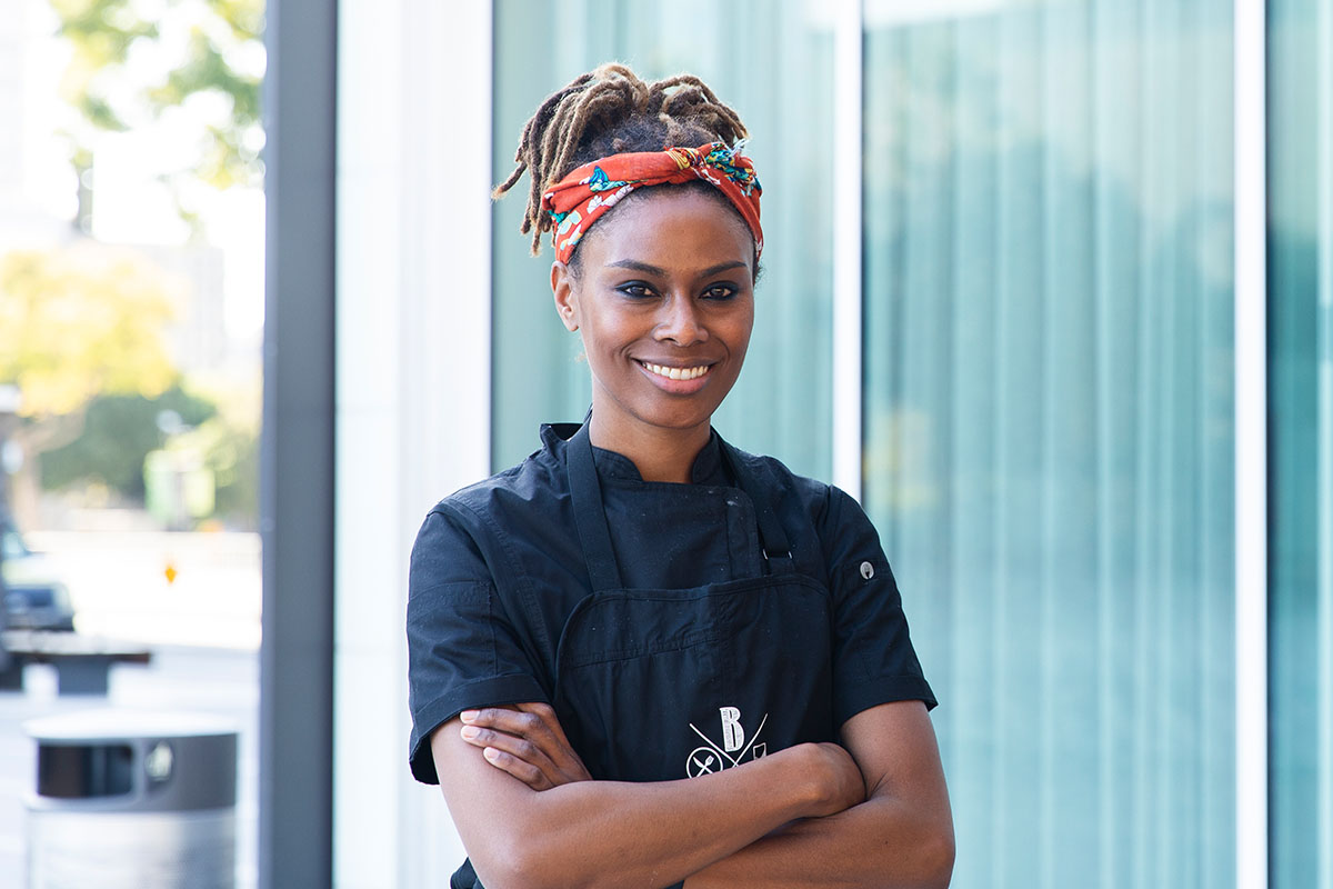 Smiling chef in a black uniform and red headband, standing with arms crossed. She is outdoors in front of a modern building, exuding confidence and warmth