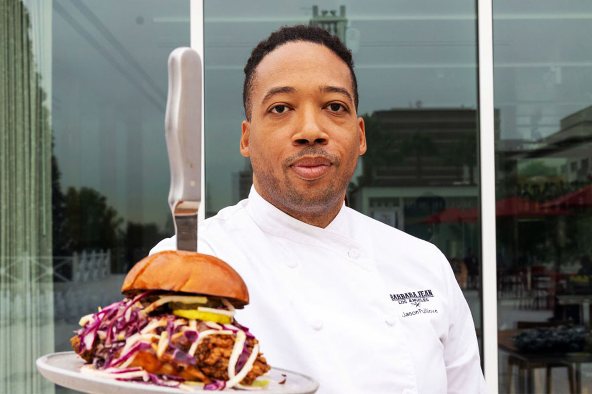 Chef in a white uniform holds a gourmet burger pierced by a knife, with colorful slaw, outside a modern glass building. The mood is professional and vibrant