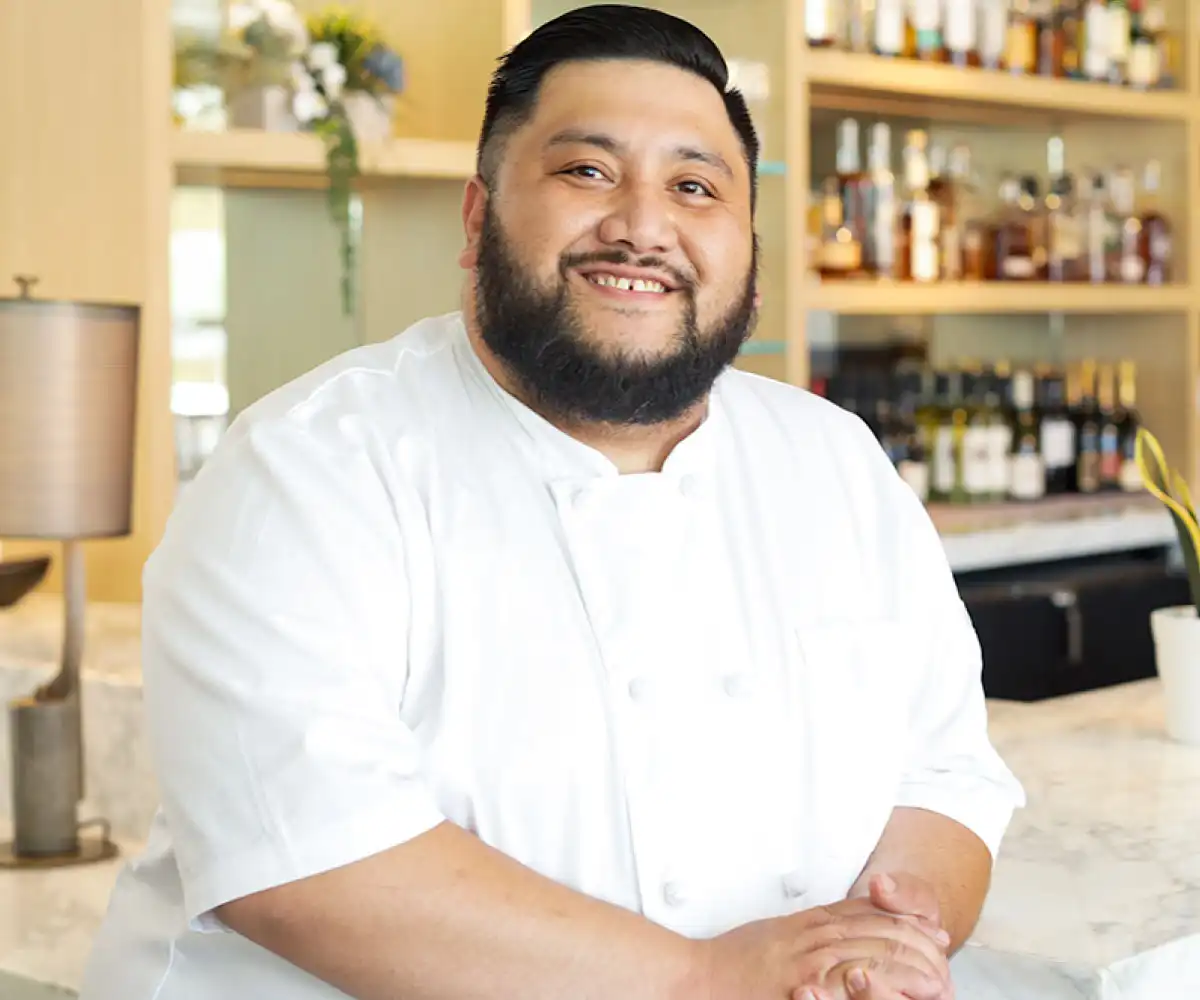 A smiling chef in a white uniform stands in a modern kitchen. Shelves of bottles are blurred in the background, creating a warm and inviting atmosphere