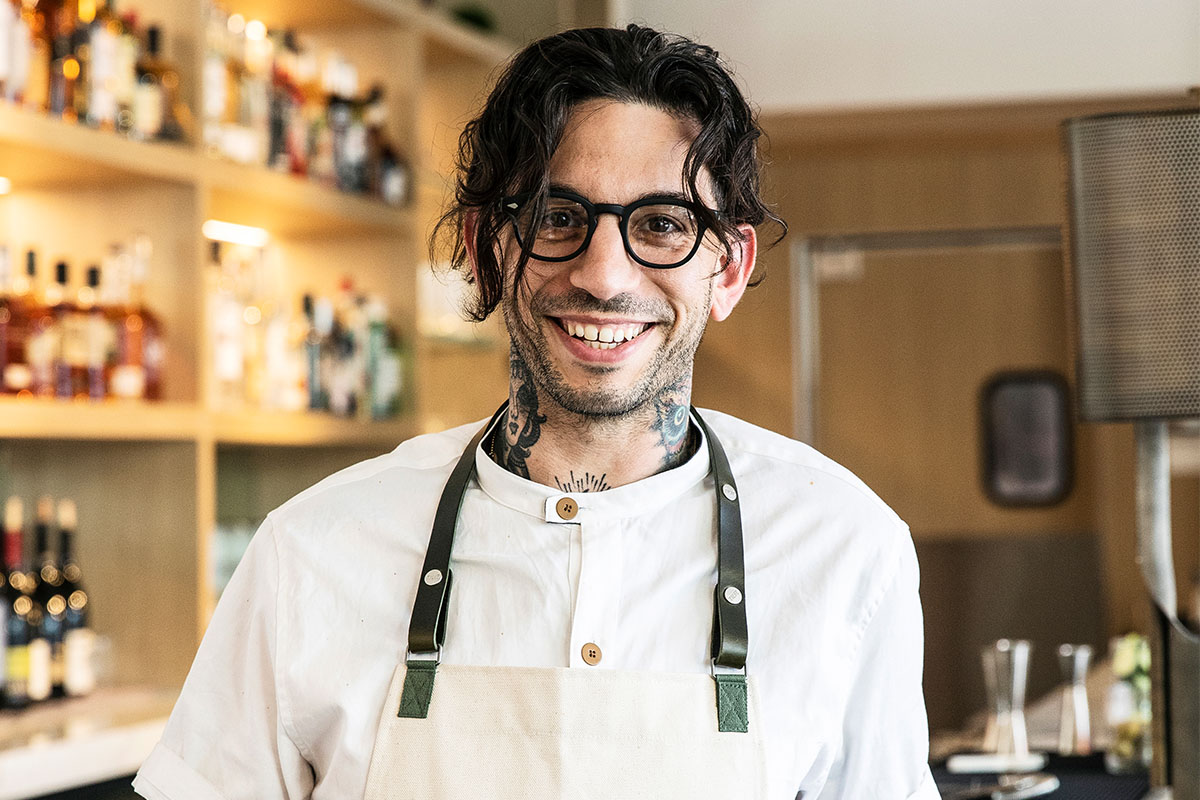 A smiling chef in a white uniform stands in a modern kitchen. Shelves of bottles are blurred in the background, creating a warm and inviting atmosphere.