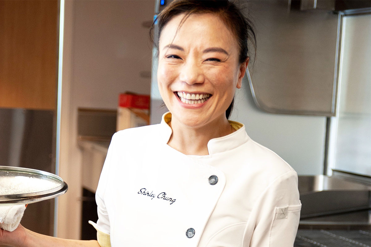 Smiling chef in a white uniform holds a silver tray in a professional kitchen, conveying warmth and hospitality. Kitchen equipment is visible in the background