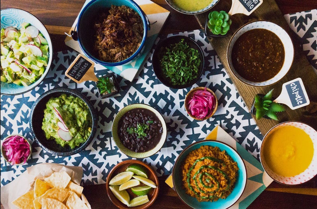 Various dishes including guacamole bowls, salad, meat, rice, sauces, fried tortillas, onions, cilantro, and lemons on a table