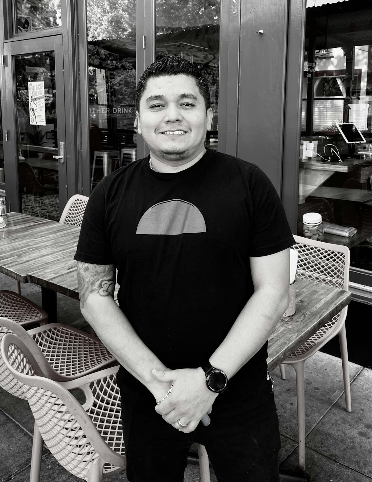 Black-shirted man posing for the camera, with chairs and tables visible in the background