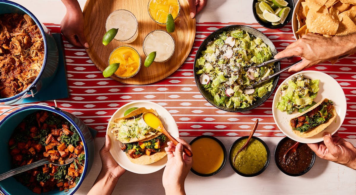 A person preparing tacos in front of a table with sauces, drinks garnished with lemons, and a bowl of salad