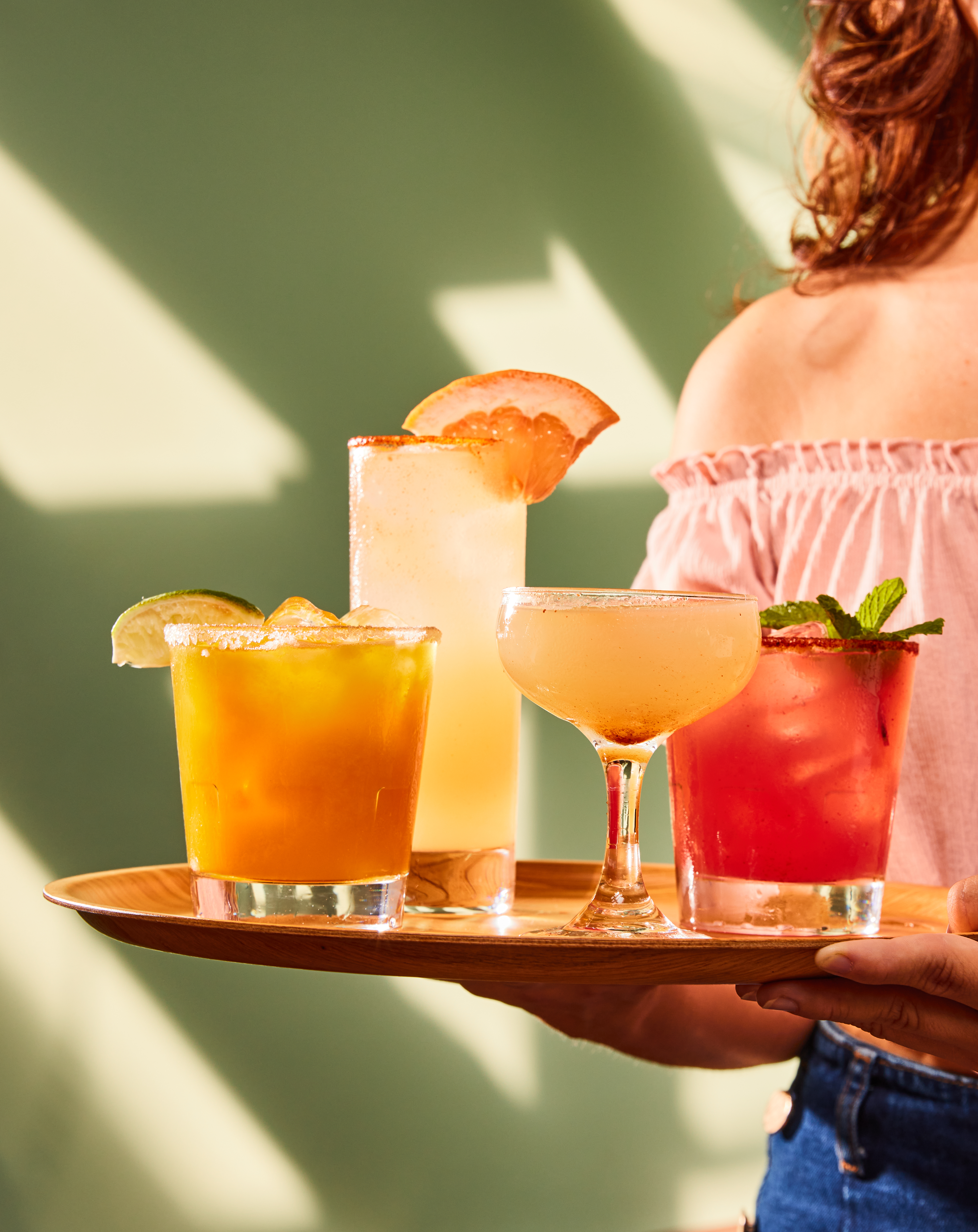 A person holding a tray of vibrant cocktails in various glasses