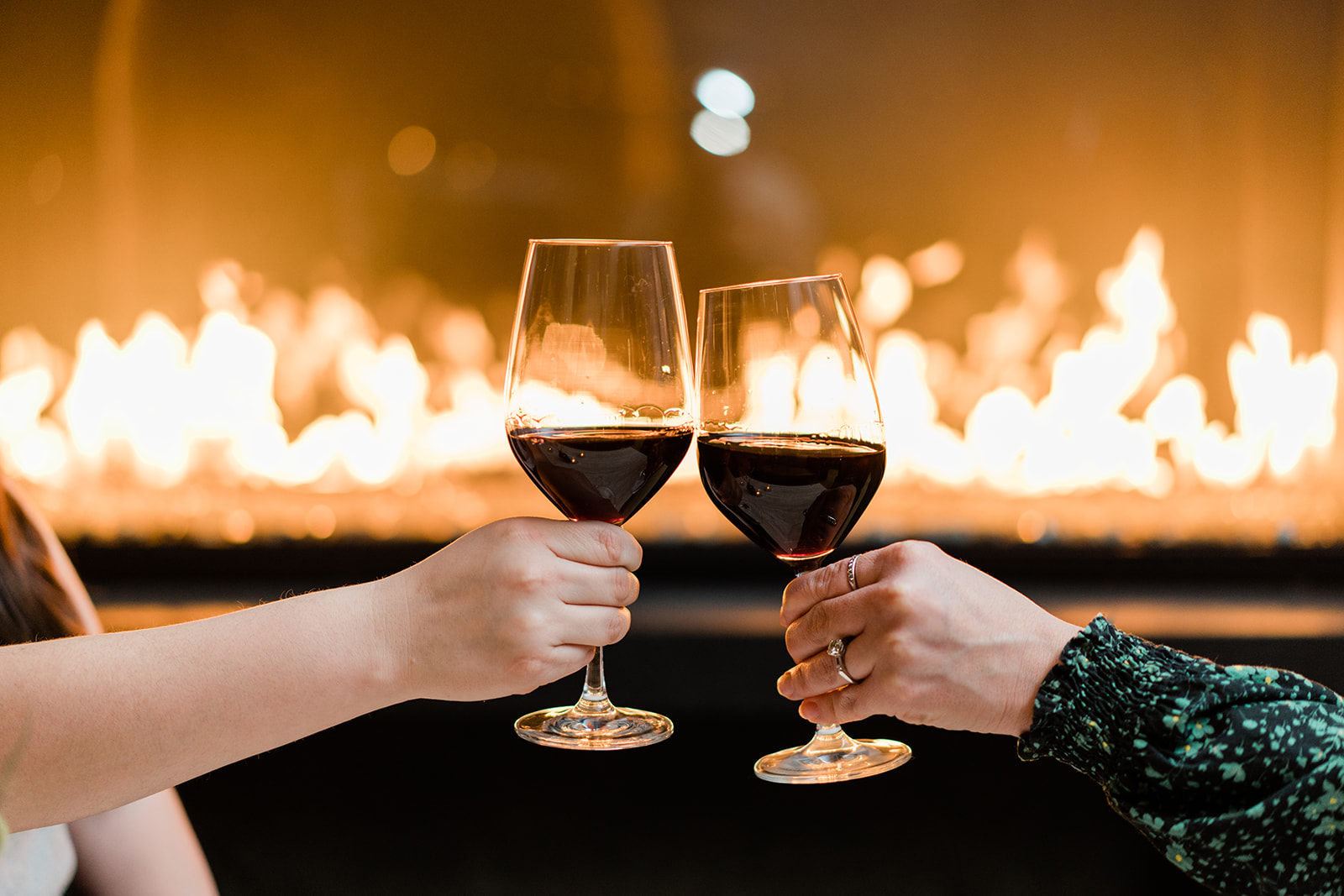 two hands clink red wine glasses together in front of a roaring fireplace