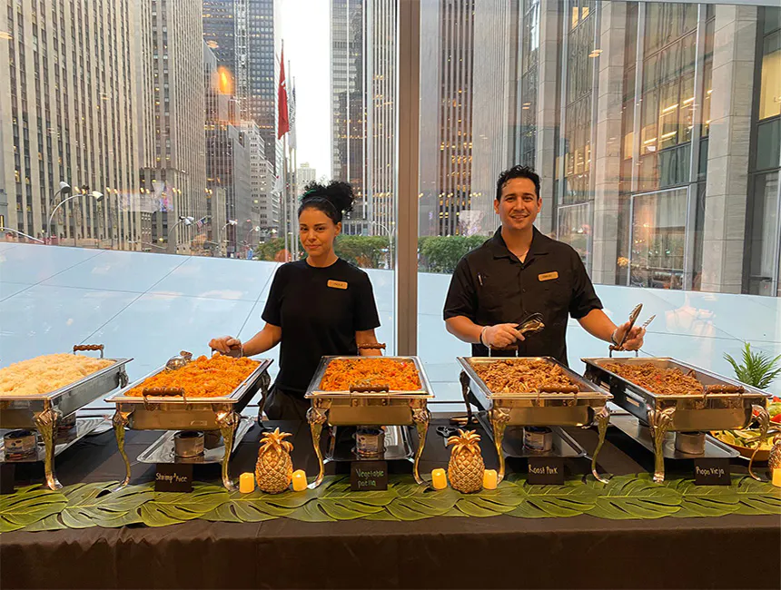Two of our Union Square, Manhattan business event caterers serving from trays of Vegetable Paella, Roast Pork, and Ropa Vieja.