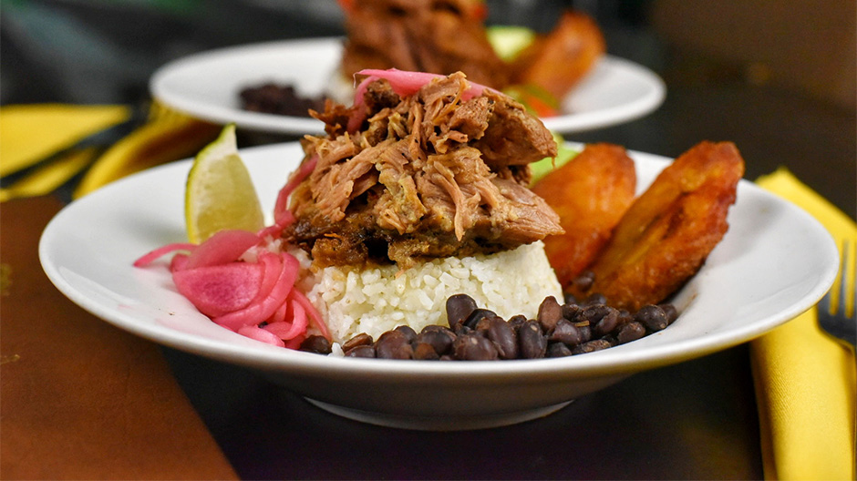 Our Pernil Lunch Bowl near Lower East Side, Manhattan, including black beans, fried plantains, and a lime.