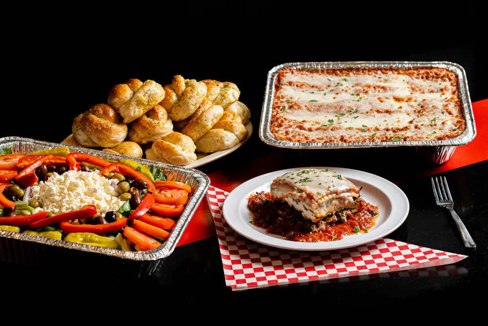 A plate of lasagna and trays of lasagna, greek salad, and garlic rolls available for Italian catering in Atlanta
