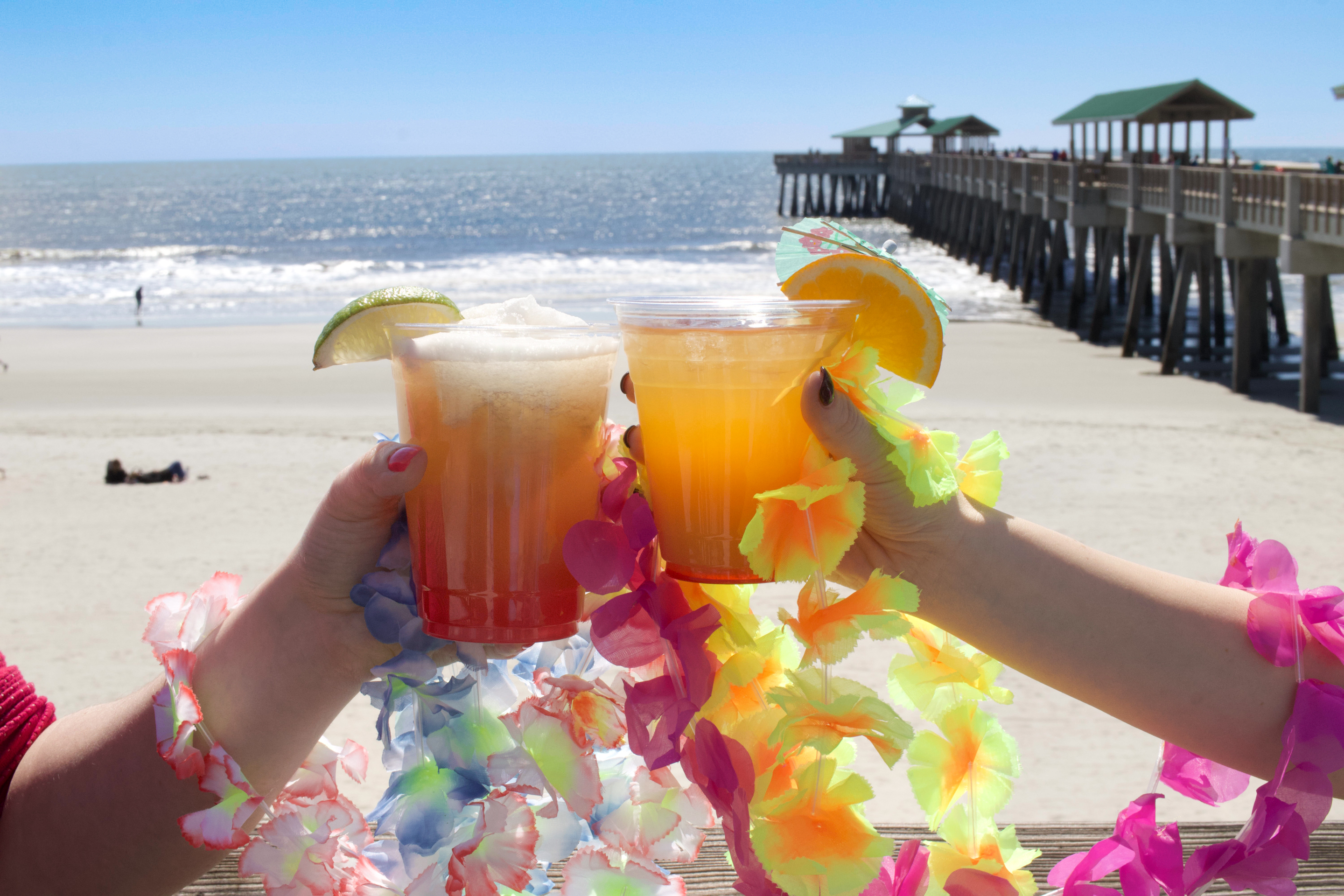 a couple of hands holding drinks with a beach and a pier in the background