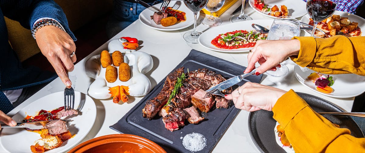 a table of food, with a person cutting steak.
