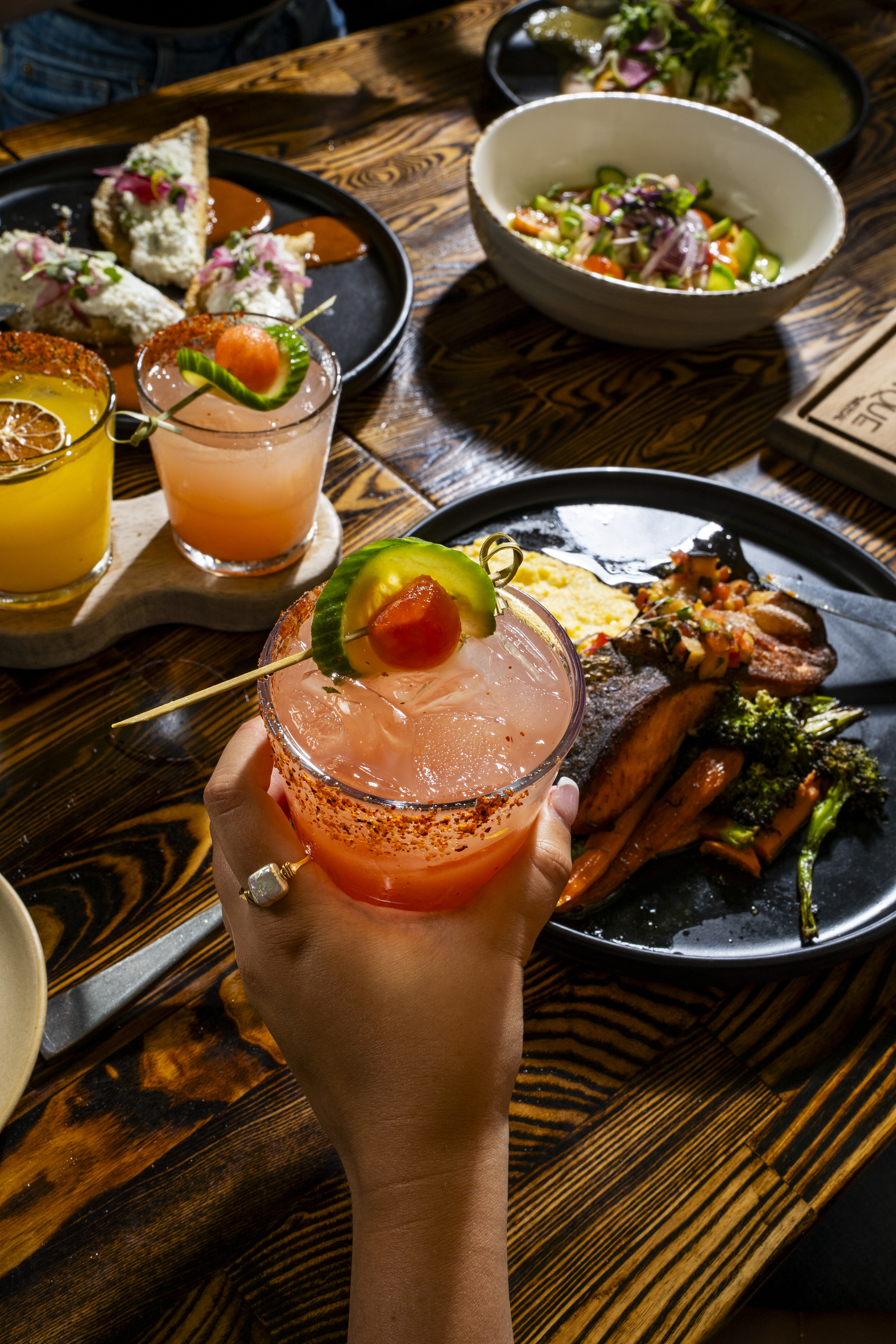 a hand holding a drink with a vegetable garnish on top of a plate of food
