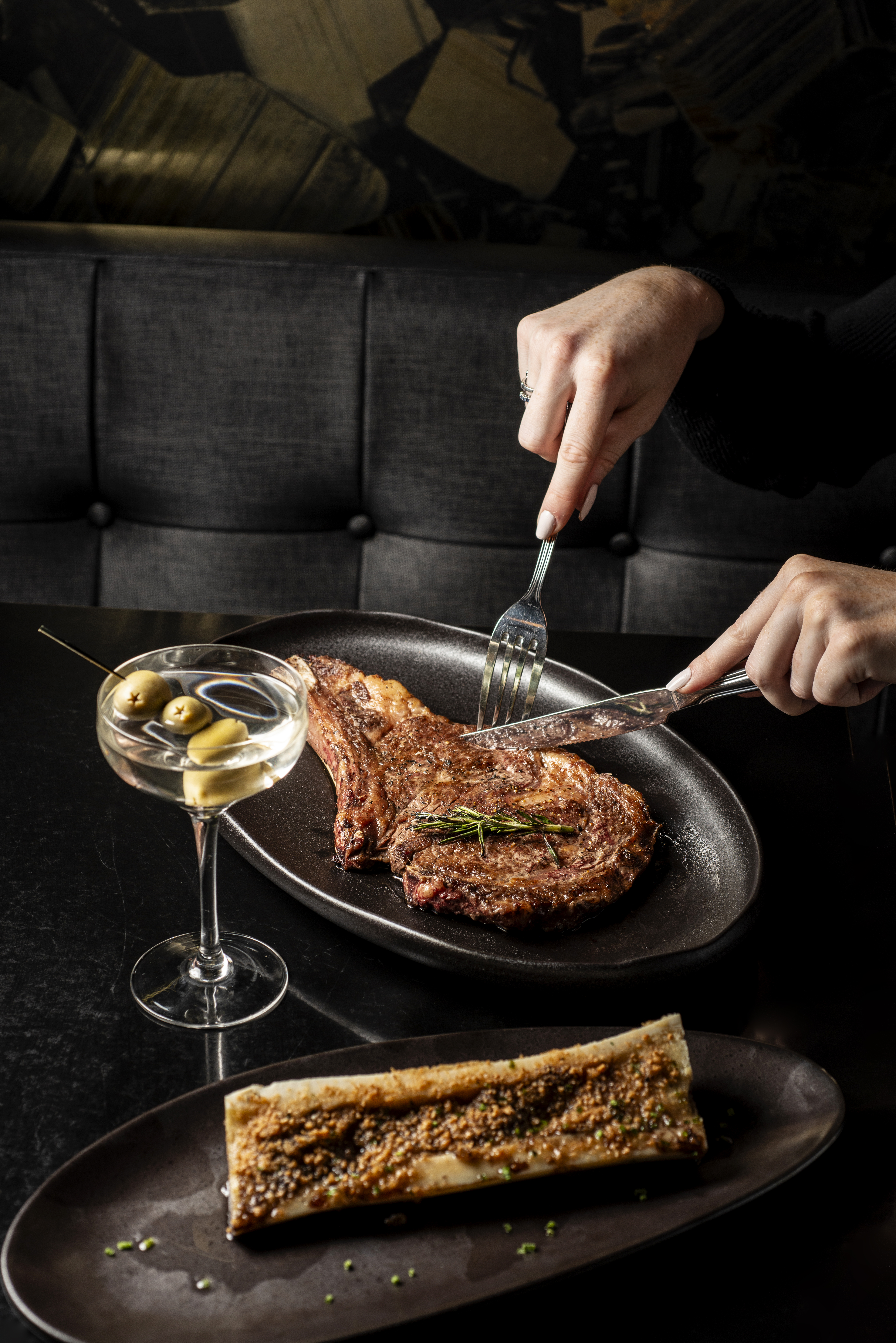 a person cutting a steak on a plate with a glass of martini