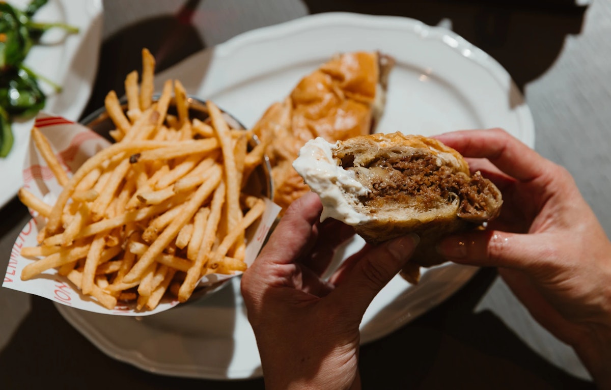 Hand holding a juicy burger with fries served on a white plate.