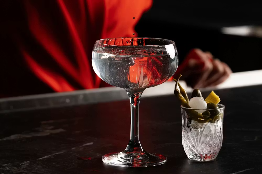 Beverage Director Amy Racine wearing a red blazer strains a clear cocktail into a coupe glass, with olives and a lemon twist.