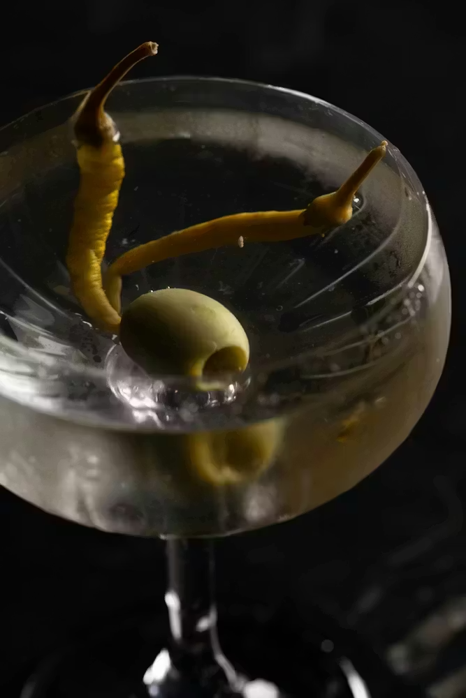 Close-up of a martini in a coupe glass garnished with a green olive and curled lemon peel, lit against a dark background.