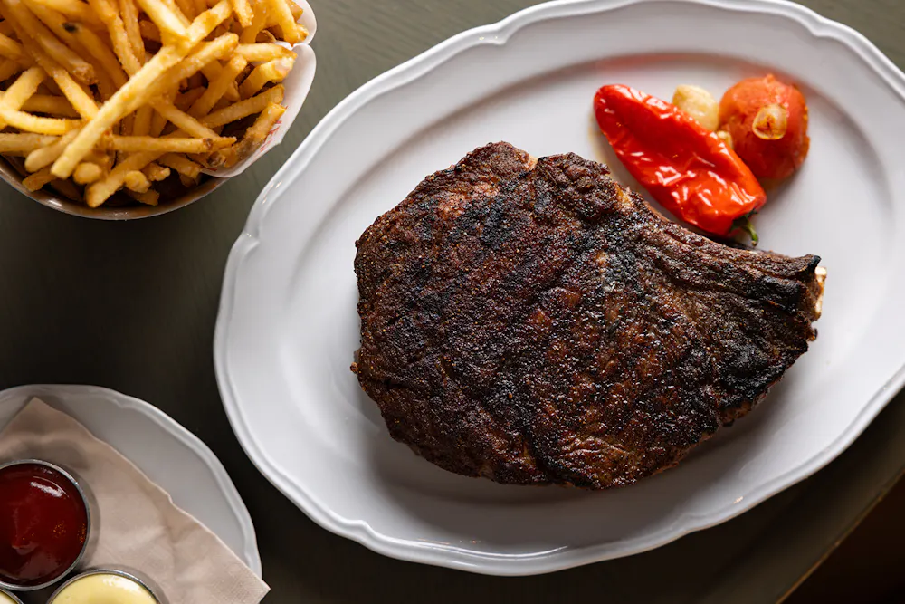 Prime 850 dry-aged steak on a white plate with roasted peppers, served with fries and dipping sauces on the side.