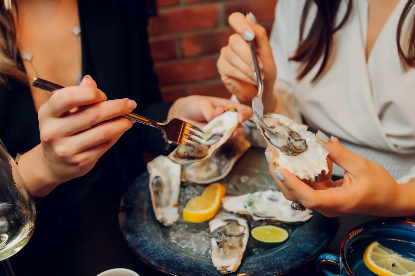 Two people enjoying fresh oysters from the half shell with lemon and lime wedges at a restaurant.