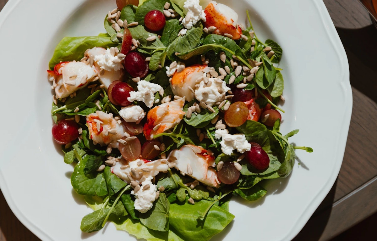 Fresh salad with lobster, grapes, goat cheese, and sunflower seeds on a white plate.