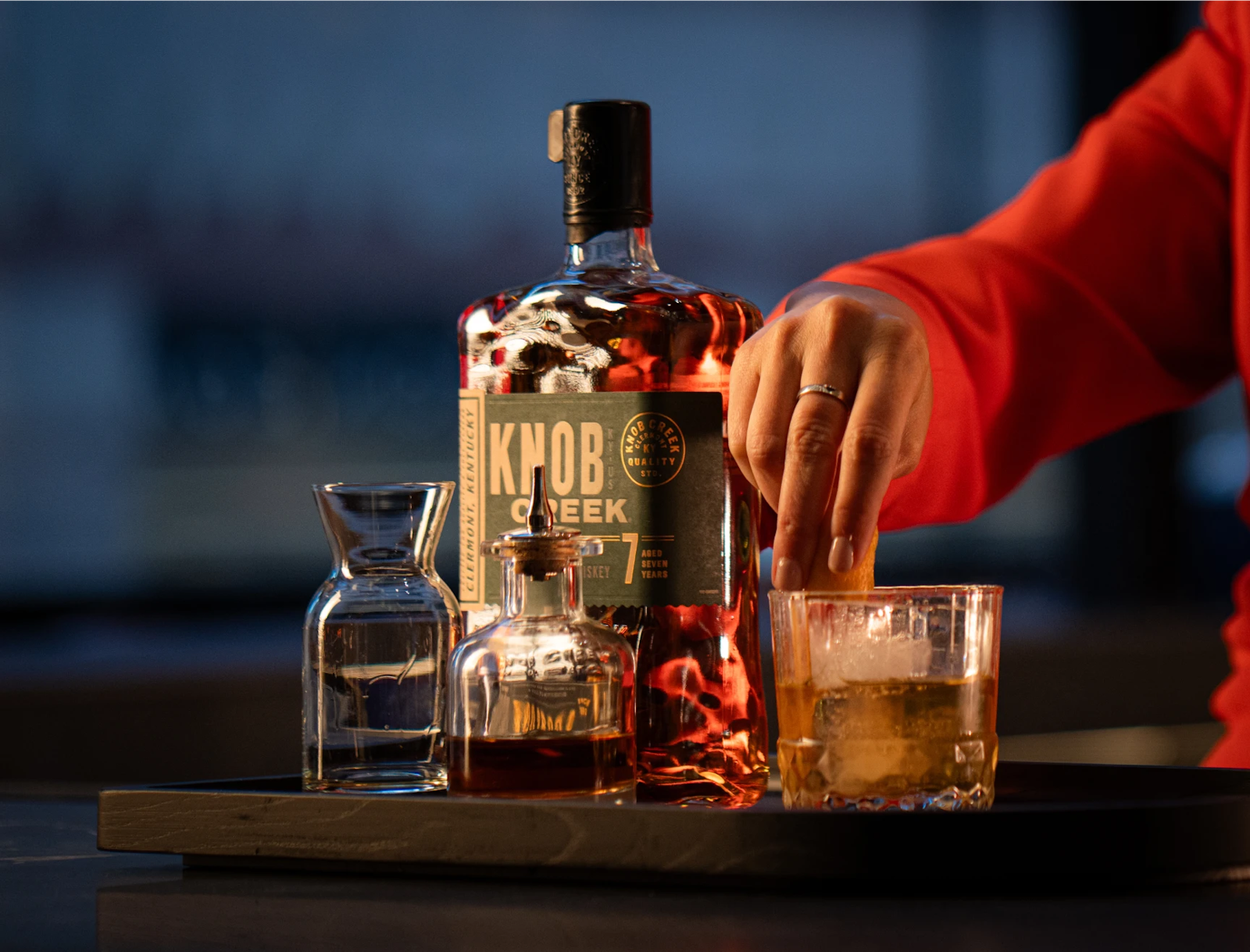 Hand preparing a whiskey cocktail with a bottle of Knob Creek bourbon and glassware on a bar tray.