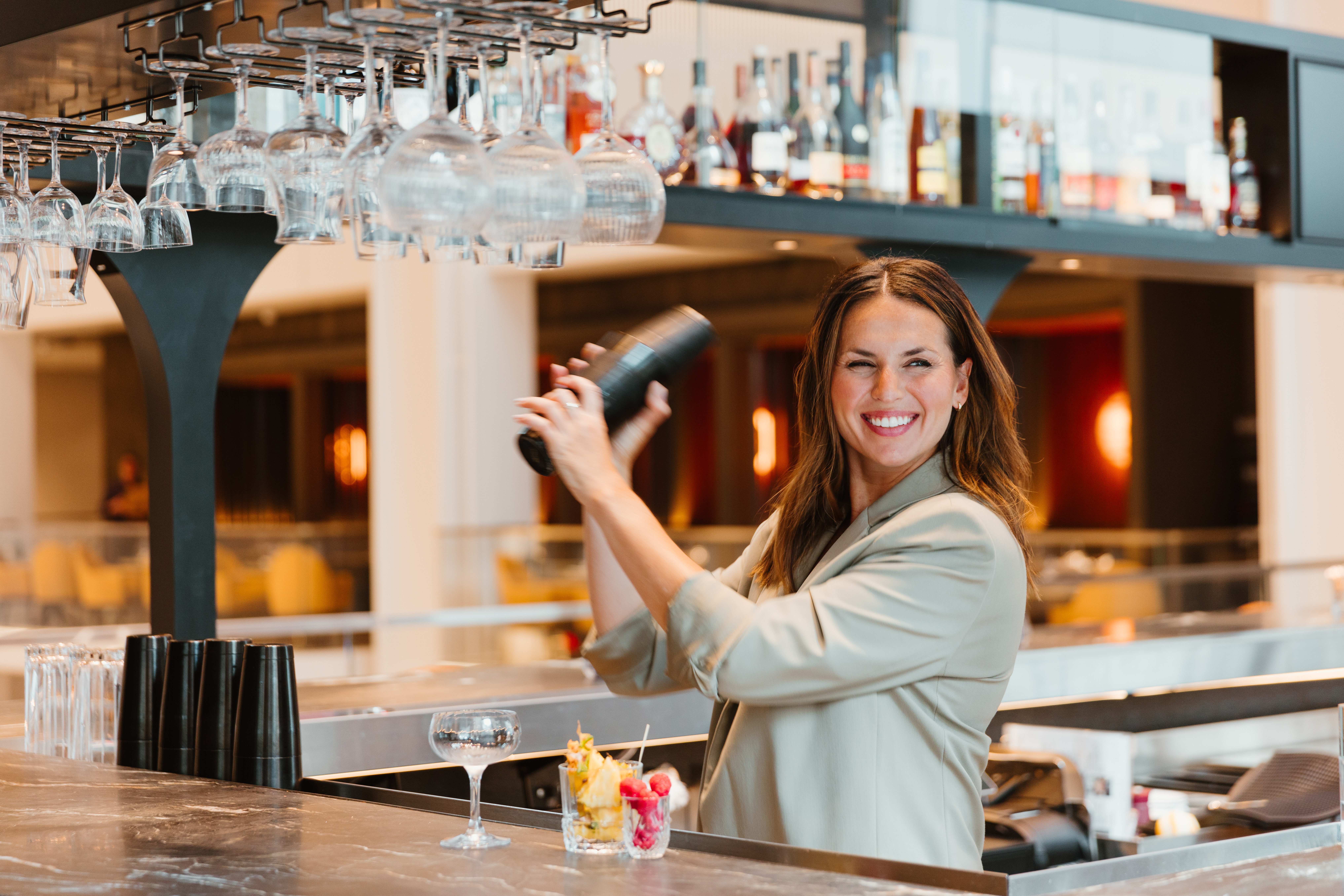Beverage Director Amy Racine smiling while shaking a cocktail behind a stylish bar with warm ambient lighting