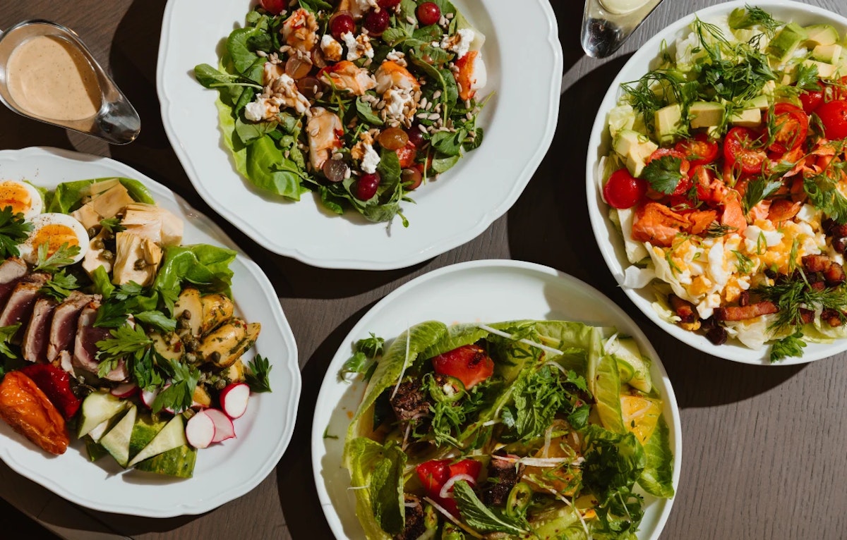 Assorted fresh salads with vibrant greens, vegetables, and proteins served on white plates.