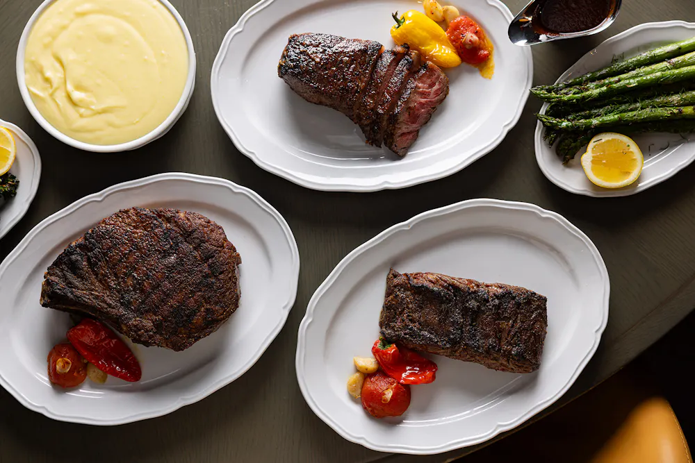 Prime 850 dry-aged steaks plated with roasted peppers and garlic, alongside asparagus, mashed potatoes, and sauce.