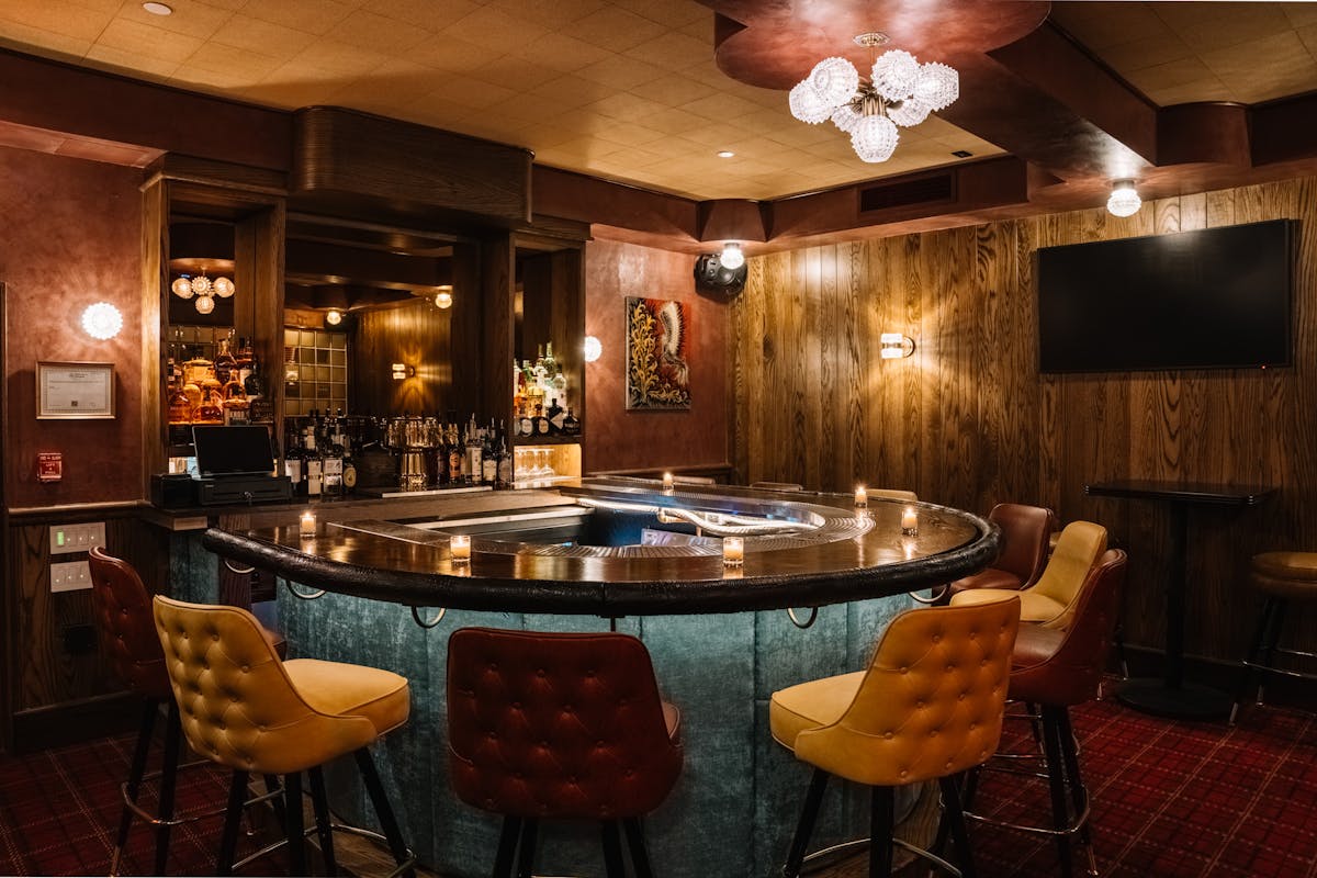 Cozy, dimly lit bar with a circular counter, colorful tufted bar stools, and warm wood-paneled walls