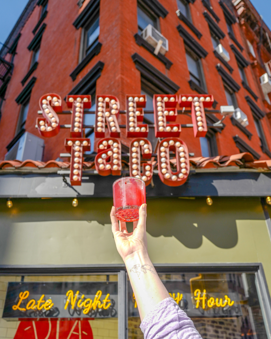 A cocktail raised in front of the Street Taco sign on Third Avenue in Midtown Manhattan, a popular taco and margarita restaurant in Kips Bay NYC