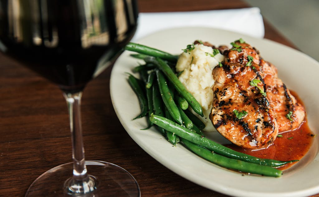 A view of plated plum pork with green beans, mashed potatoes and a glass of red wine on a wooden table.