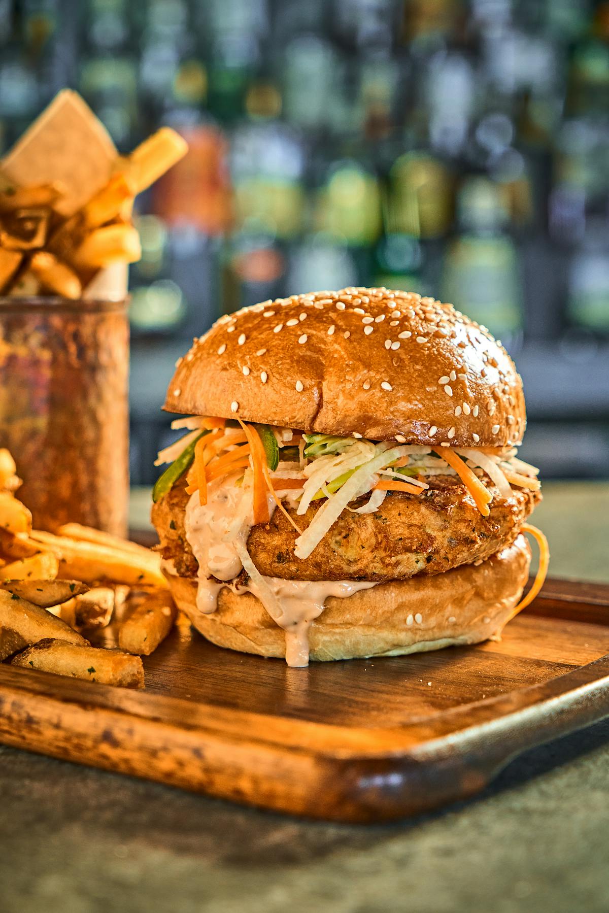 a saucy fried chicken sandwich on a wooden tray next to seasoned french fries 