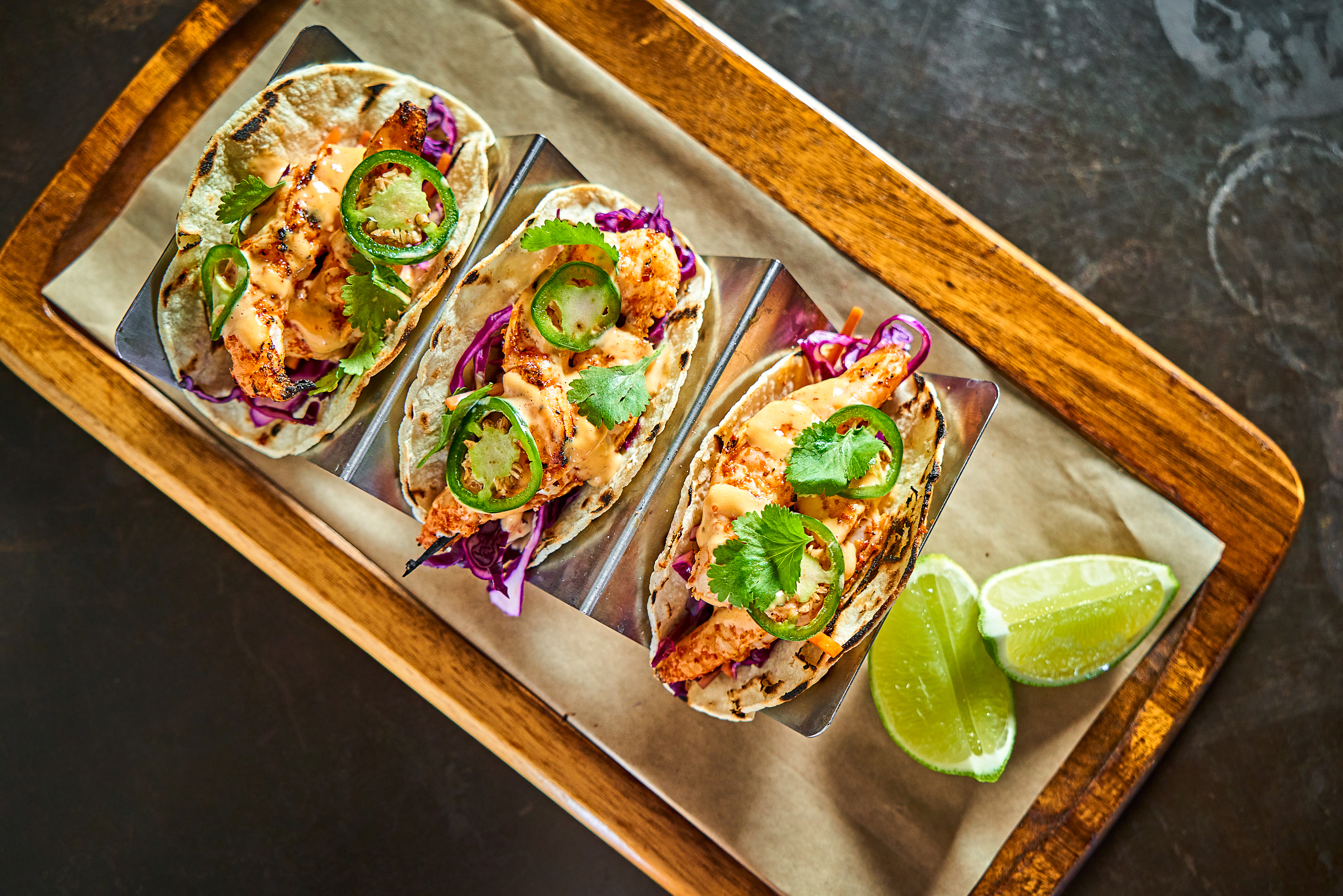 three spicy chicken tacos with fresh jalapeno and lime slices on the side on a wooden tray